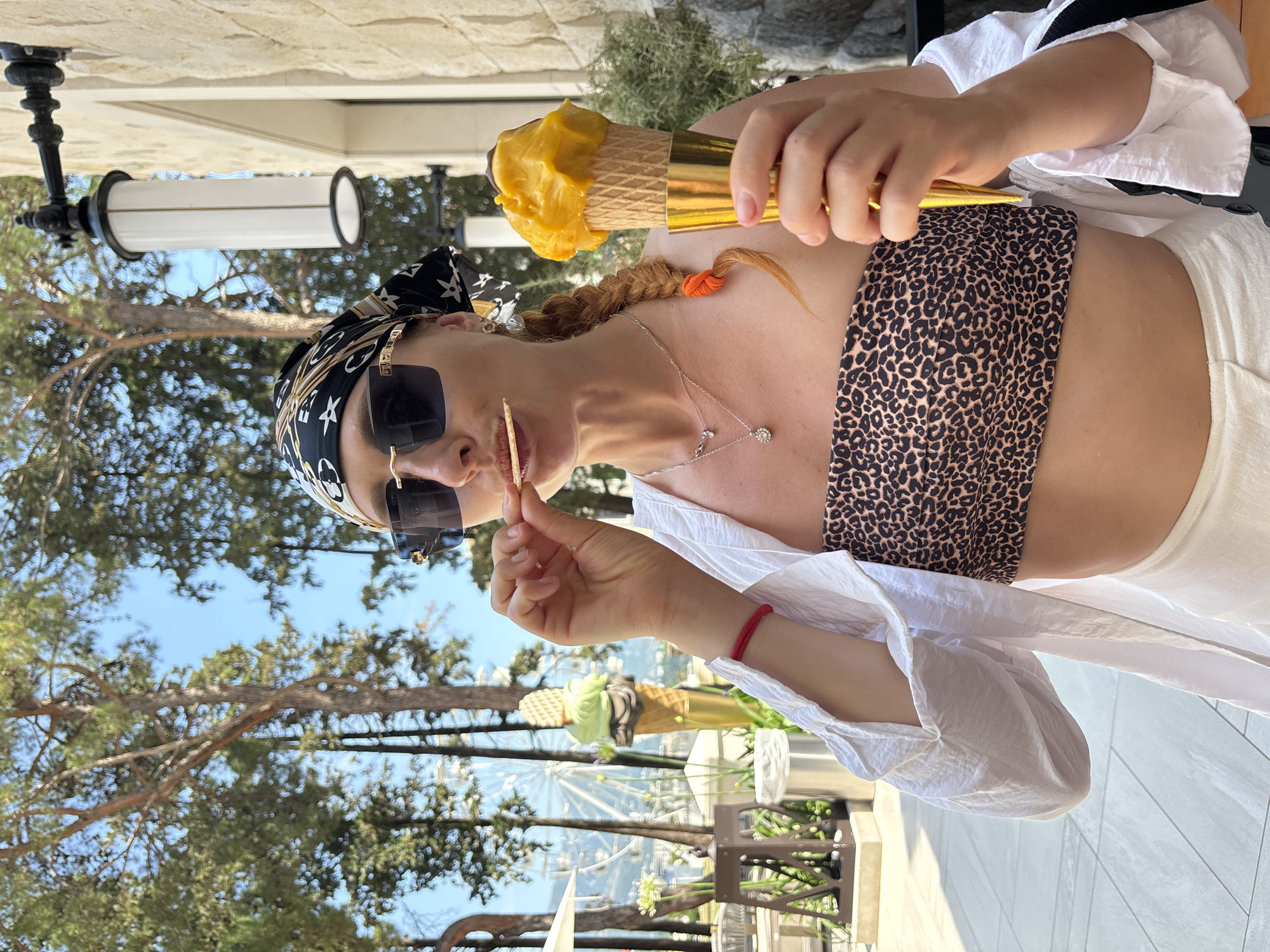 In Budva, Montenegro, a woman with red hair styled in a braid and adorned with a bandana and sunglasses is captured in a medium shot. She wears a leopard print bandeau top and a white open shirt, with white pants partially visible. In her right hand, she holds a golden ice cream cone with yellow-orange ice cream and a dark chocolate topping. Her left hand is raised to her mouth, where she is eating a thin, light-colored cracker or chip. The background reveals a bright, sunny outdoor environment with lush green trees, a stone building with a wall lamp, and in the distance, a Ferris wheel. The overall scene suggests a casual, warm-weather setting.