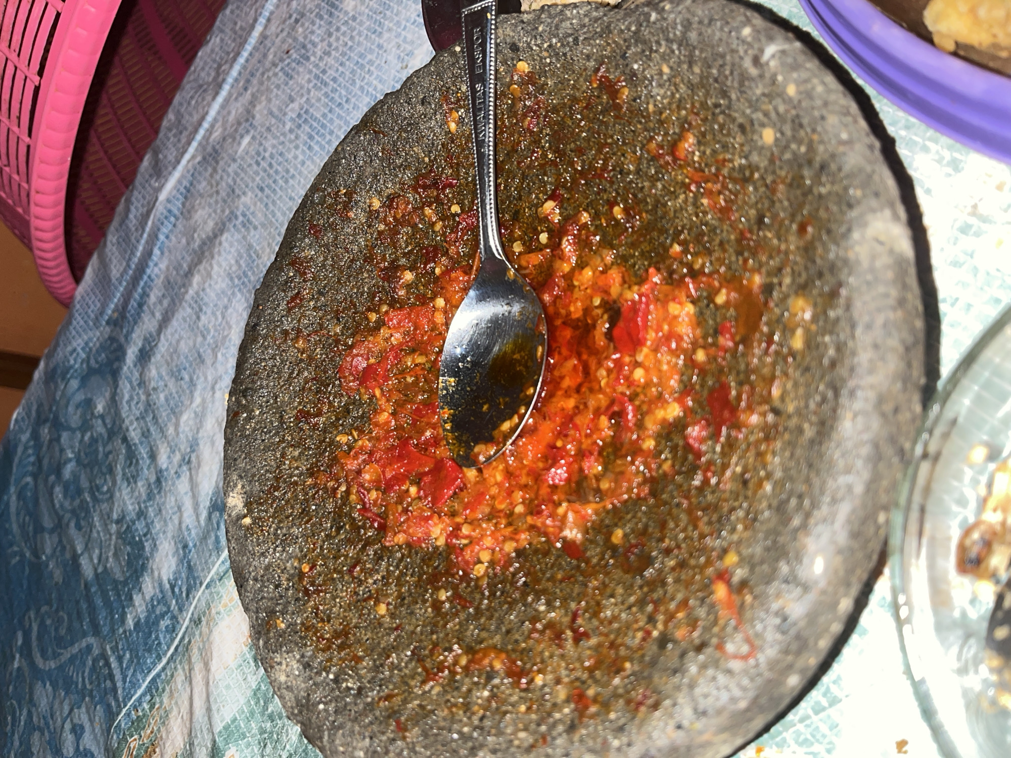 The image shows a close-up view of a mortar and pestle containing a red chili paste, likely sambal, on a patterned cloth. A silver spoon rests on the paste. The background reveals a portion of a pink basket and other blurred objects. The scene is set in Glagah Kulon, Indonesia.