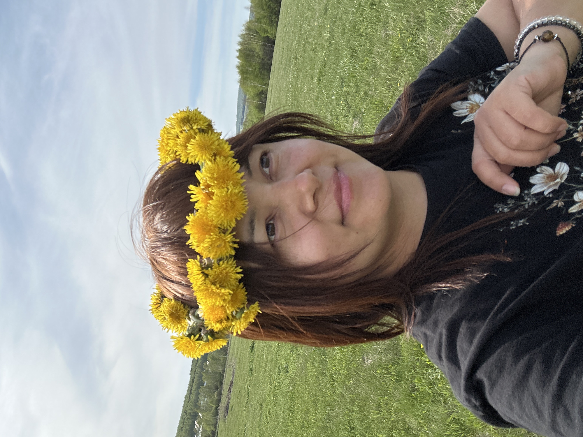 A woman is posing for a selfie outside in a field of green grass with a tree line in the background. She is wearing a crown of yellow dandelions. She is wearing a dark colored shirt with white flower details. On her right wrist, she is wearing two bracelets. It is daytime, and the sky is blue with light white clouds. The location appears to be in a rural area near Unknown, Unknown.