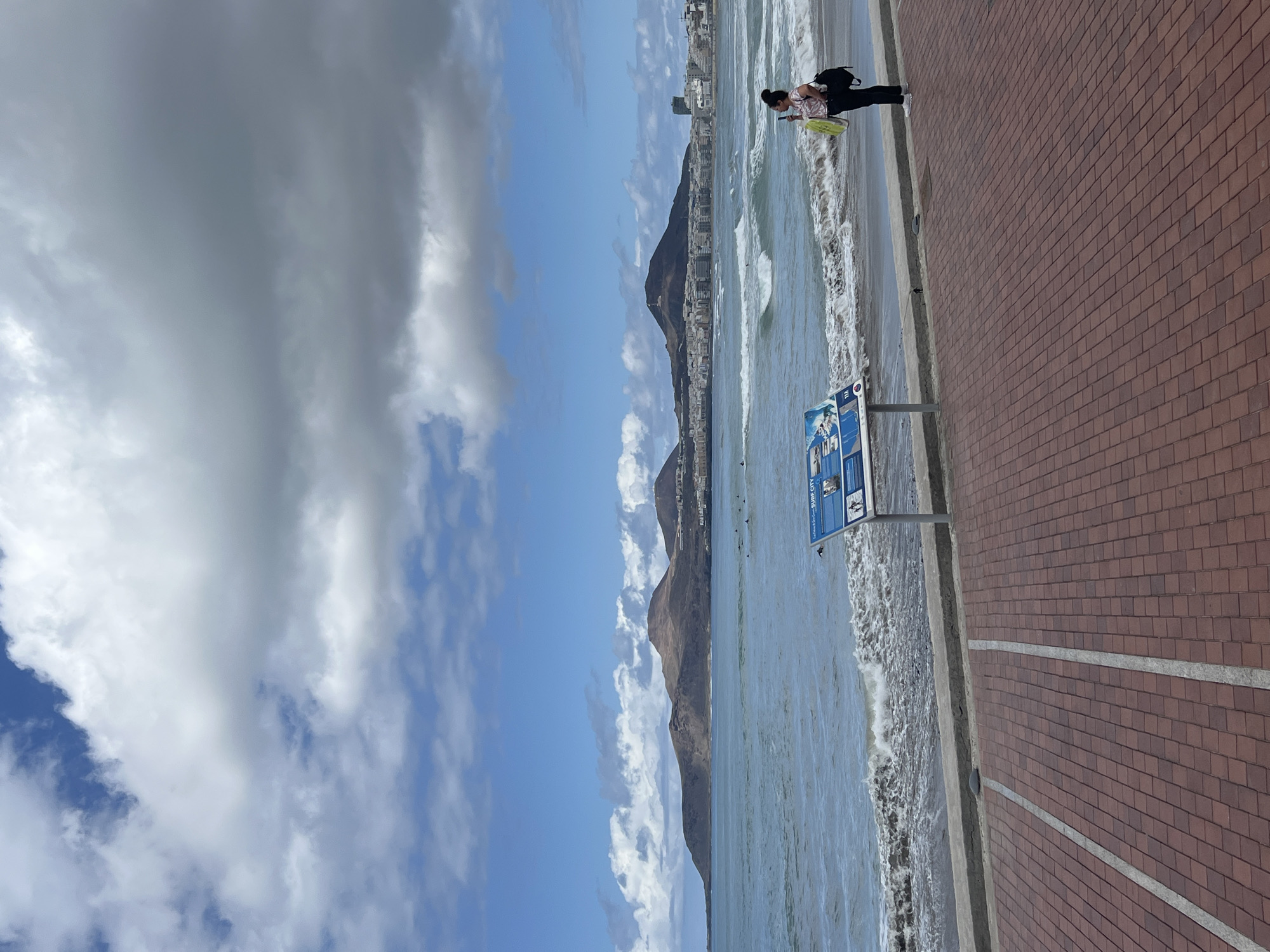 This media file captures a vibrant coastal scene in Las Palmas De Gran Canaria, Spain, on what appears to be a bright, partly cloudy day. The image is oriented vertically, showing the horizon running from left to right.
In the foreground, a wide promenade paved with reddish-brown bricks stretches across the lower right of the frame, separated from the beach by a concrete curb. A person, possibly an adult female, with dark hair tied back, stands on the edge of this promenade near the water. She is wearing a light-colored, possibly patterned, short-sleeved top, dark trousers, and white athletic shoes. She carries a dark backpack and a bright yellow bag. Her gaze appears to be directed towards the ocean.
To the left of the person, positioned on the promenade, is a blue rectangular informational sign mounted on a grey pole. The prominent text "Surf City" is clearly visible, along with smaller text and pictograms below it, likely providing details about surfing or beach regulations.
The midground features the Atlantic Ocean with moderate waves breaking on a sandy shore. Dark shapes in the water suggest the presence of several surfers. Beyond the immediate coastline, the background reveals a lengthy stretch of a developed city with numerous buildings clustered along the coast, culminating in a series of large, dark mountains rising majestically.
The sky above is a dynamic mix of clear blue patches and large, white and grey cumulus clouds, indicating fair but somewhat overcast weather conditions. The overall impression is one of a bustling yet scenic coastal environment, with activities like surfing taking place under a changing sky.