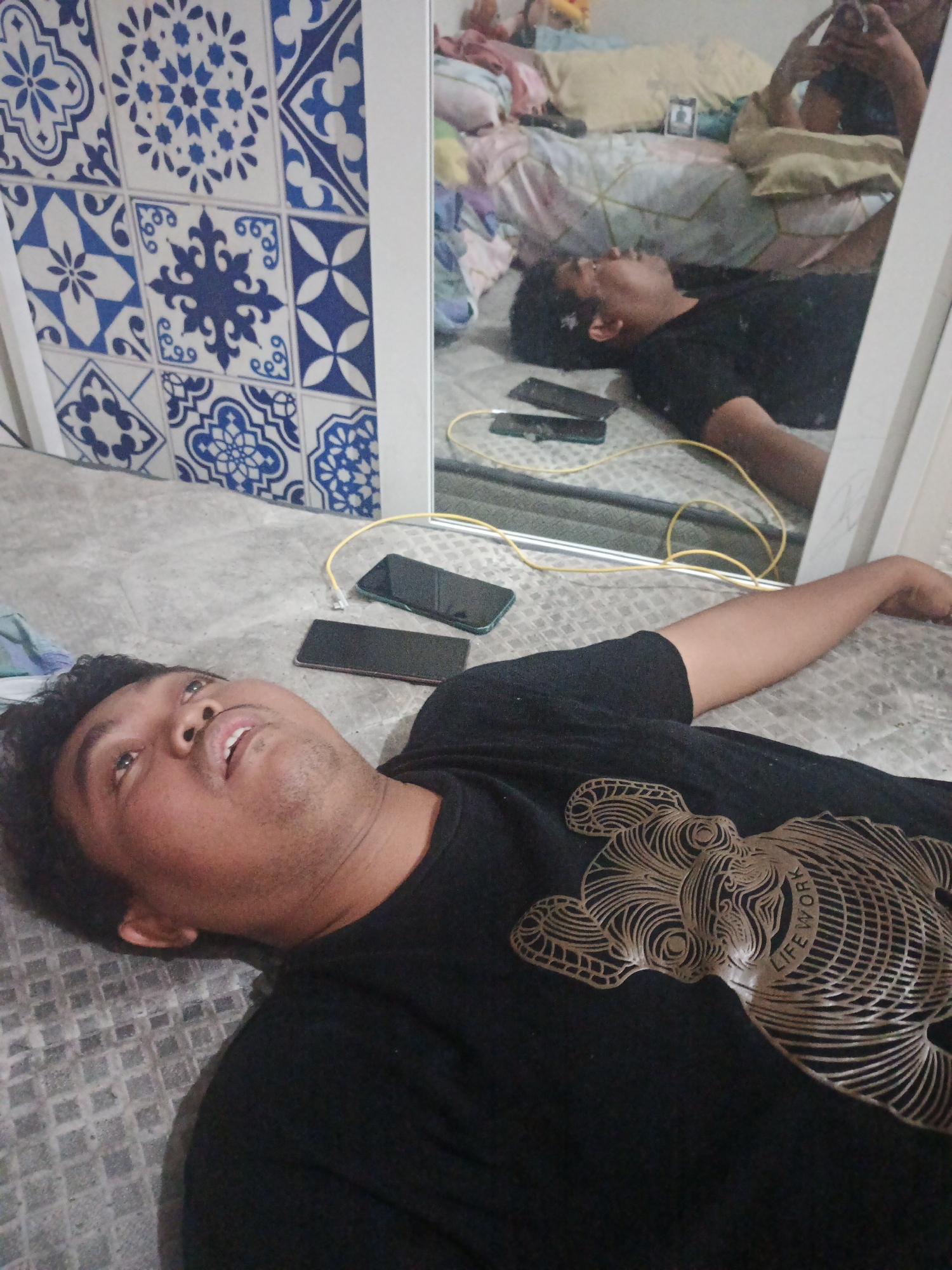 A man is lying on his back on a textured surface, possibly a bed or mattress, with his head turned to his left and eyes open. He is wearing a black t-shirt with a gold line-art graphic of what appears to be an animal. His right arm is outstretched to his side. Three mobile phones are on the surface near him: two are black, and one is green. A yellow cable, possibly a charger, is also present. In the background, a mirror reflects the man and the room behind him. The mirror shows him from a different angle, and behind him, another person is visible in the reflection, holding a mobile phone. The room appears cluttered with bedding and pillows. To the left of the main subject, a wall or decorative panel with a blue and white patterned tile design is visible. The scene is indoors.