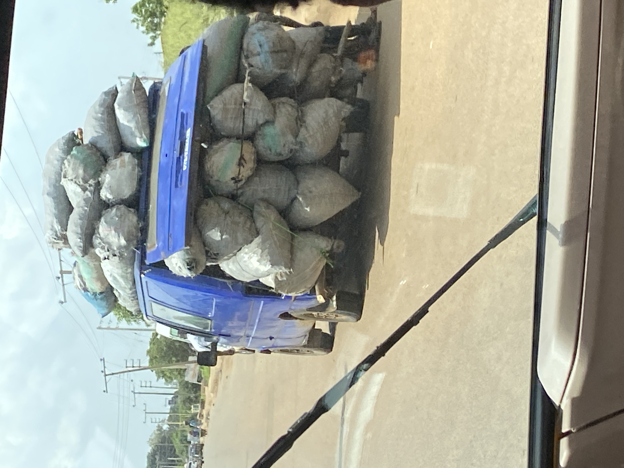 A blue van is heavily laden with large, bulging sacks of goods, some of which appear to be charcoal or agricultural produce. The sacks are piled high on the roof and sides of the van, secured with ropes. The van is traveling on a paved road, with a few other vehicles and pedestrians visible in the background. Overhead power lines are visible in the distance, along with trees and a cloudy sky. The image appears to be taken from inside another vehicle, with part of a windshield wiper in the foreground. The scene is likely located in Bukuru, Nigeria, a town known for its mining and agricultural activities.