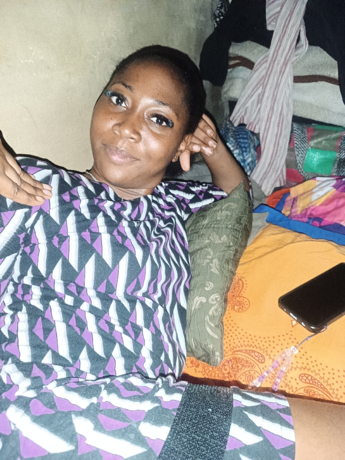 A woman is lying on a bed, resting her head on her hand. She wears a geometric patterned dress with purple, white, and gray triangles. A green pillow is beside her, and a smartphone rests on the orange patterned bedspread. In the background, clothes are piled up. The woman is looking at the camera with a slight smile and has long eyelashes.