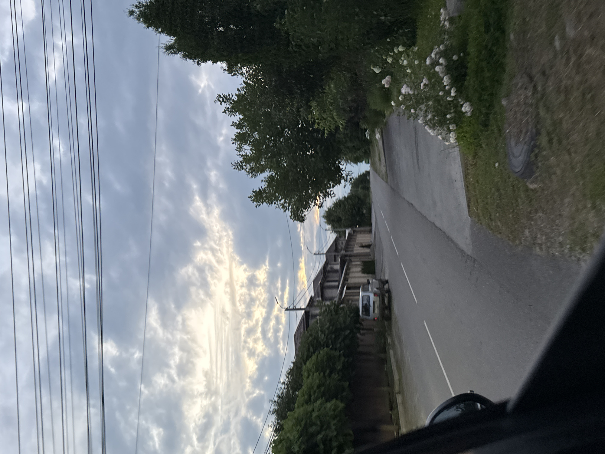 The media file captures a scene from a vehicle's perspective, looking out onto a road in Mikhaylovsk, Russia. The sky is a prominent feature, filled with a dramatic, cloudy pattern, suggesting either dawn or dusk due to the soft, diffused light. The clouds appear textured and illuminated, with hints of yellow and orange indicating the sun's position, although the sun itself is not directly visible.
The road, paved and marked with white lines, curves gently to the right. A small, silver van is visible on the road, moving away from the viewer. On the left side of the road, a row of houses or buildings can be seen, with dark, pitched roofs and some visible windows. Adjacent to the buildings is a dense line of trees, providing a green, natural border. Further to the right, a grassy area is visible, with a circular manhole cover partially obscured by debris. White flowers bloom along a hedge bordering the road.
Several power lines are strung across the sky on the left side of the frame, running parallel to each other and creating a graphic element against the cloudy backdrop. The overall mood is somewhat serene, with the natural elements of the sky and greenery contrasting with the man-made structures and the road. The lighting suggests a time of day when natural light is soft and atmospheric, possibly early morning or late evening. No people are clearly visible.