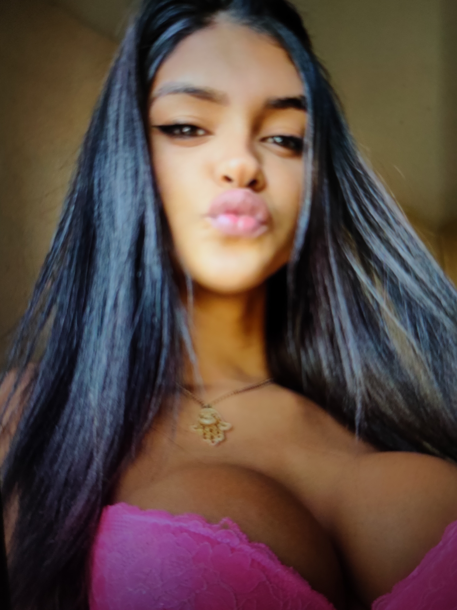 A close-up, head-and-shoulders shot captures a young woman with long, dark hair and dark eyes. She is looking directly at the camera with her lips pursed. She is wearing a pink lacy bra and a gold necklace with a hamsa pendant. The background is a blurred beige wall. The lighting suggests an indoor setting, possibly with natural light coming from the side.