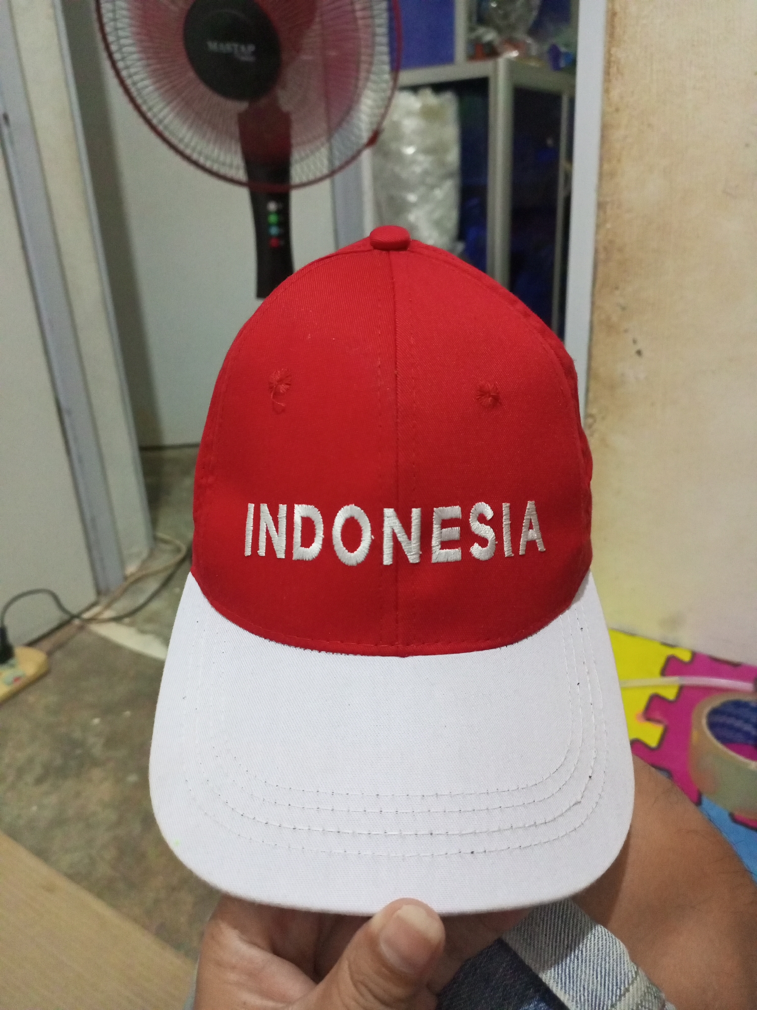 A red and white baseball cap with the word "INDONESIA" embroidered in white on the red portion is held in a person's hand. The cap is two-toned, with the front section red and the brim white. The background includes a standing fan, a wall, and some colorful puzzle pieces on the floor. The scene appears to be indoors in an unspecified location.