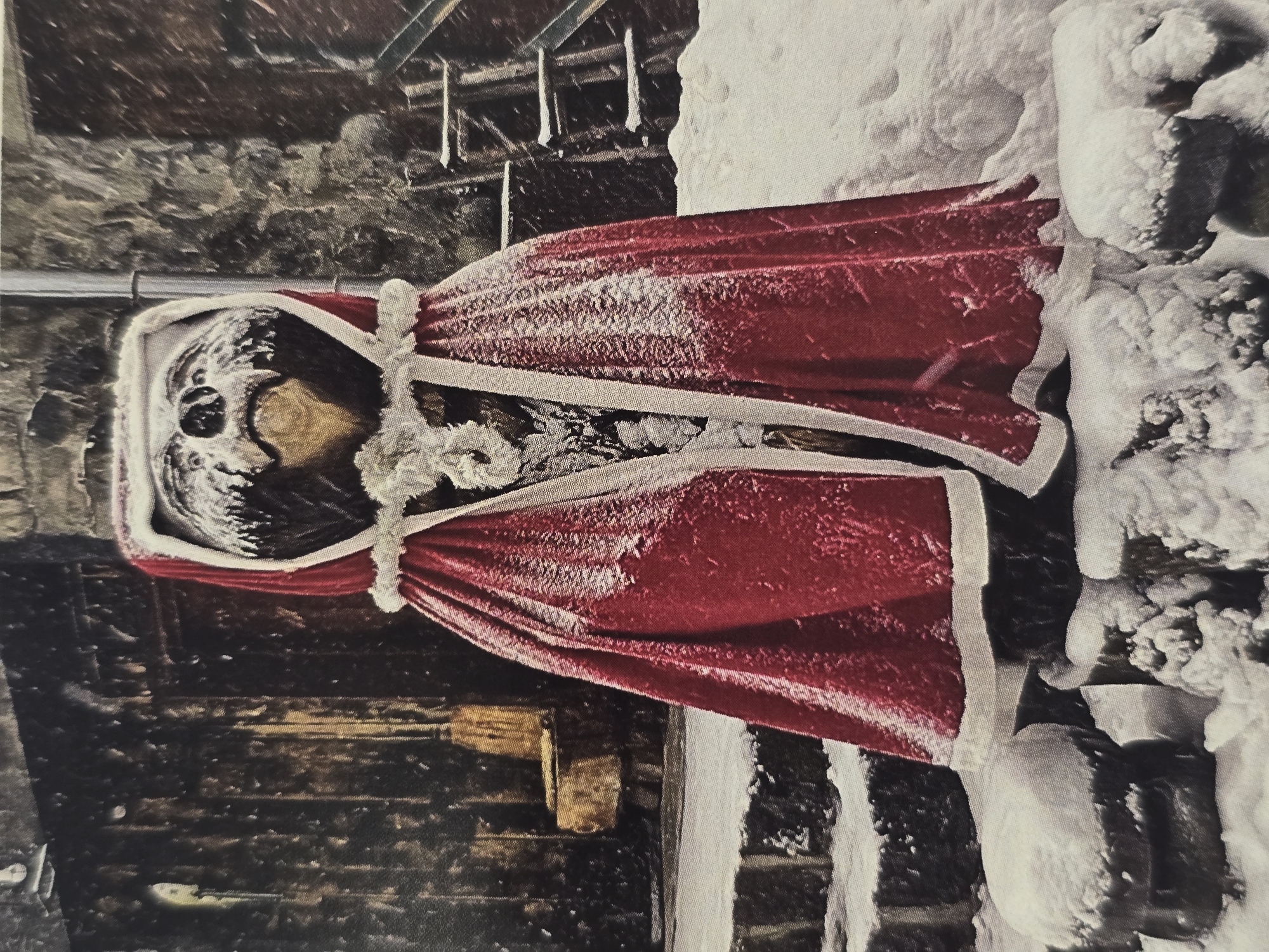 The image depicts a Christmas decoration of a figure in a Santa Claus-style cloak covered in snow. The cloak is red with white trim. The figure is positioned next to a snow-covered surface. The background appears to be a building facade, possibly constructed of brick or stone, with some wooden structures overhead. The photograph was likely taken in Sofia, Bulgaria.
