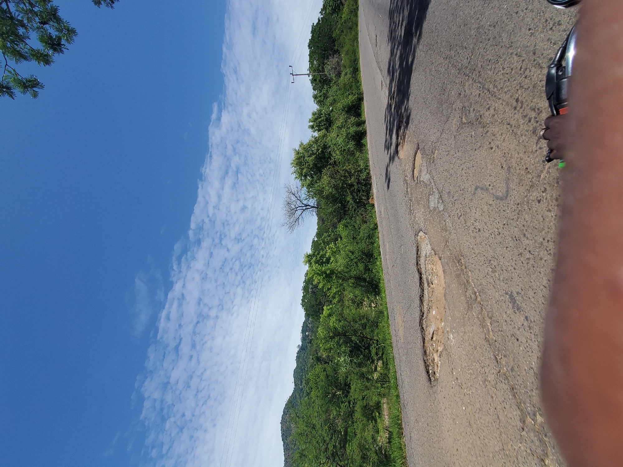 The image depicts a road with several potholes, suggesting poor road conditions. The road curves to the right, leading into a landscape with lush green trees and hills in the background under a bright blue sky with scattered white clouds. A telephone pole with wires is visible on the right side of the image. In the foreground, a person's hand and part of a motorcycle handlebar are visible, indicating the photo was taken from a moving vehicle. The scene is likely a rural or semi-rural area, possibly on the outskirts of Gwolong, Nigeria, given the presence of natural landscape and the general appearance of the road and infrastructure. There are no other individuals or significant interactions visible in the frame.