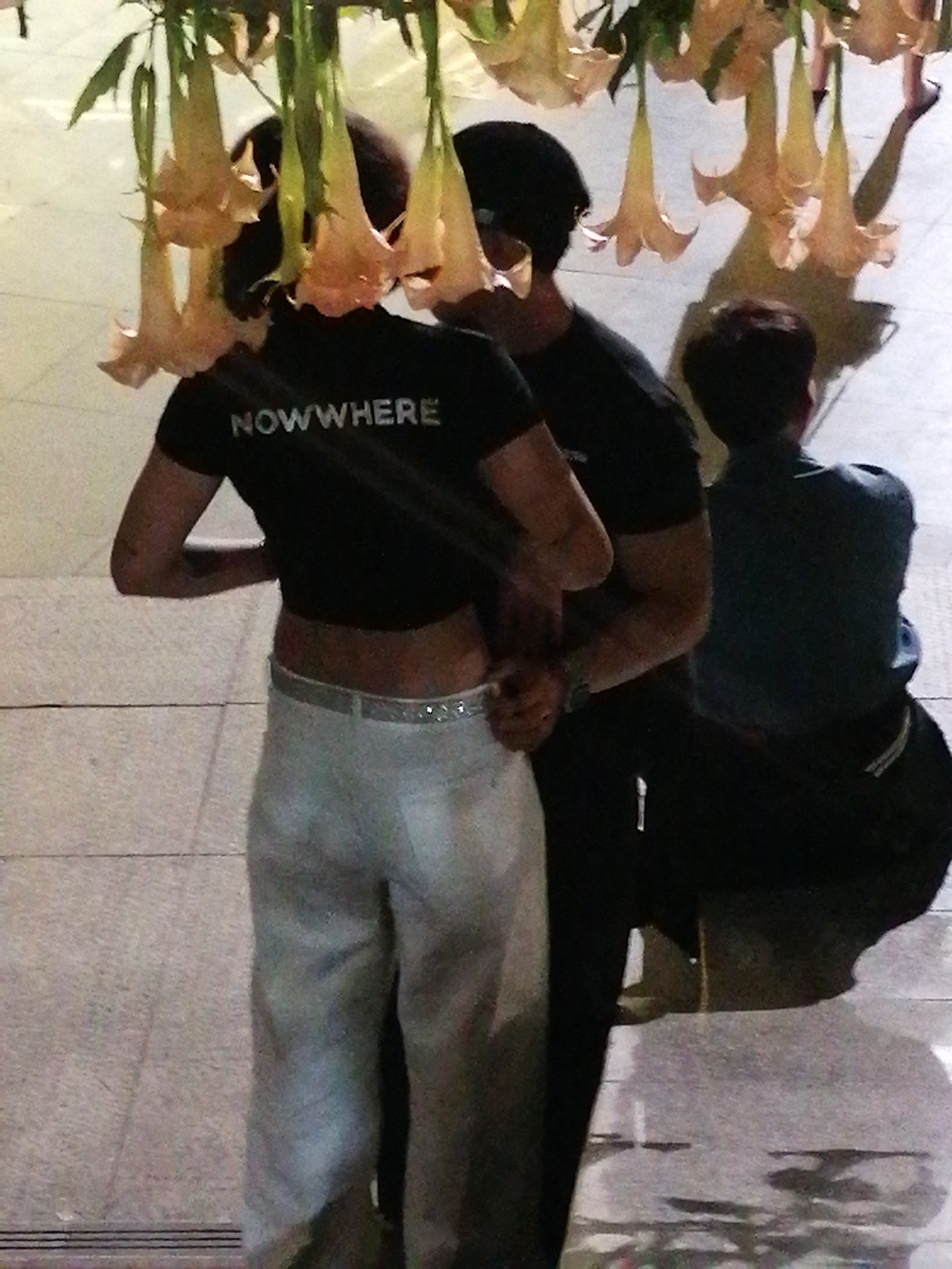 The image shows a group of people standing under hanging yellow flowers in Da Lat, Vietnam. The foreground features a woman in a black crop top with the word "NOWHERE" printed on it and light-colored pants. She is standing next to a man also wearing a black shirt. Another person is seated behind them, wearing a dark shirt. The background depicts a tiled walkway and more hanging flowers, characteristic of the floral displays in Da Lat. The overall scene appears to be outdoors in a public space.