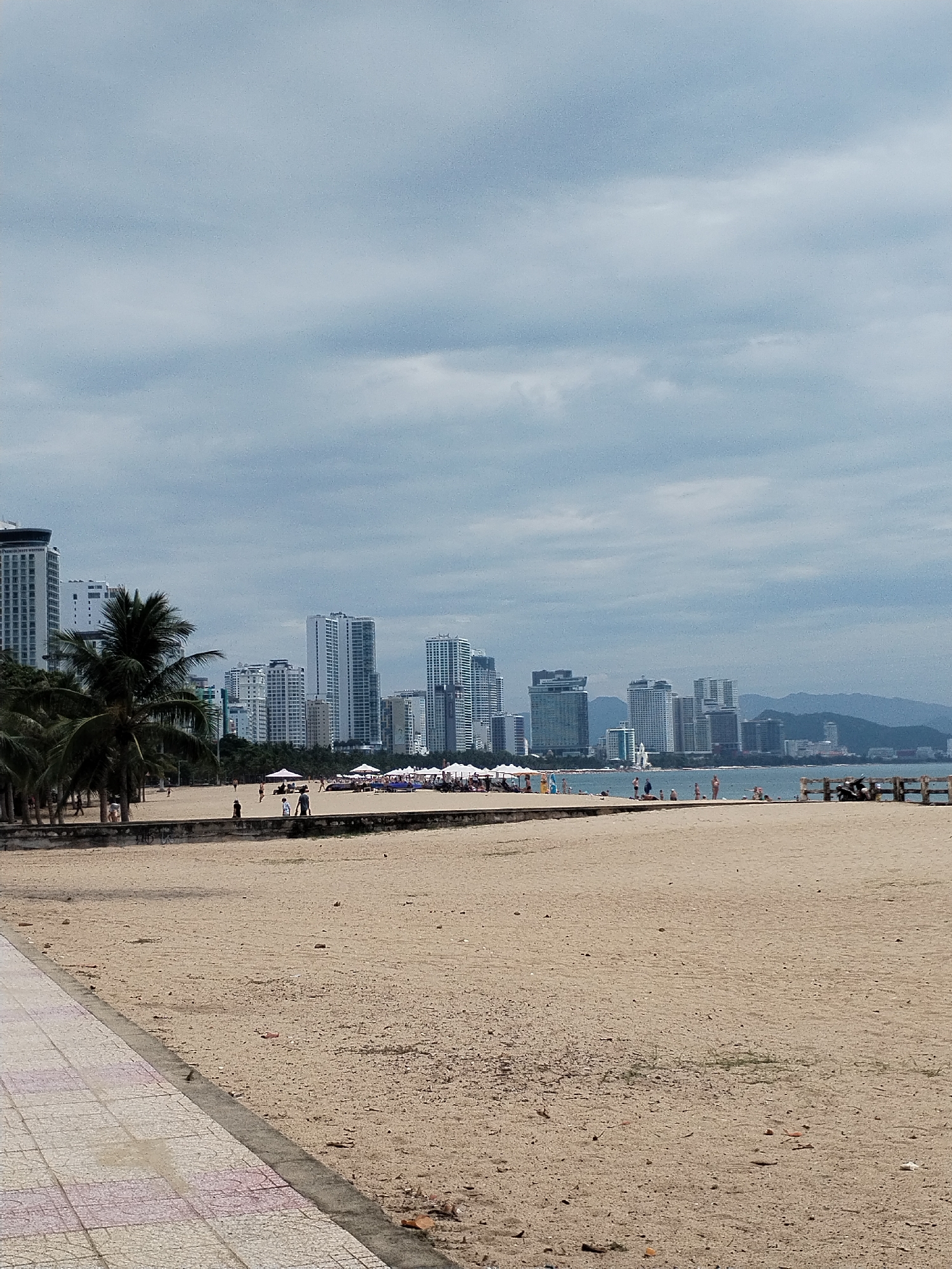 This media file presents a wide-angle view of a beach in a bustling coastal city, identified as Nha Trang, Vietnam. The scene is set under an entirely overcast sky, characterized by thick, grey clouds, indicating daytime without direct sunlight.
In the foreground, a tiled walkway runs along the left edge, bordering a broad expanse of light brown sand. A low, weathered stone wall separates this sandy area from the main beach, which extends towards the ocean. To the left, a cluster of palm trees lines the edge of the beach, partially obscuring some of the buildings.
The midground features numerous small figures of people scattered across the sandy beach. Some are seen walking, while others gather near groups of white beach umbrellas and loungers. In the distance, the ocean meets the shore, with a few individuals visible closer to the water. On the far right, a small wooden pier or barrier extends into the water, and what appear to be motorbikes are parked nearby on the sand.
The background is dominated by a dense urban skyline, featuring numerous modern high-rise buildings and skyscrapers, indicative of a developed city. Faintly visible behind the city structures and extending into the distance are rolling mountains. On the tallest building on the far left, there is some faint, illegible text at the top, possibly part of a hotel name. The overall impression is a vibrant resort city under a calm, cloudy sky.
