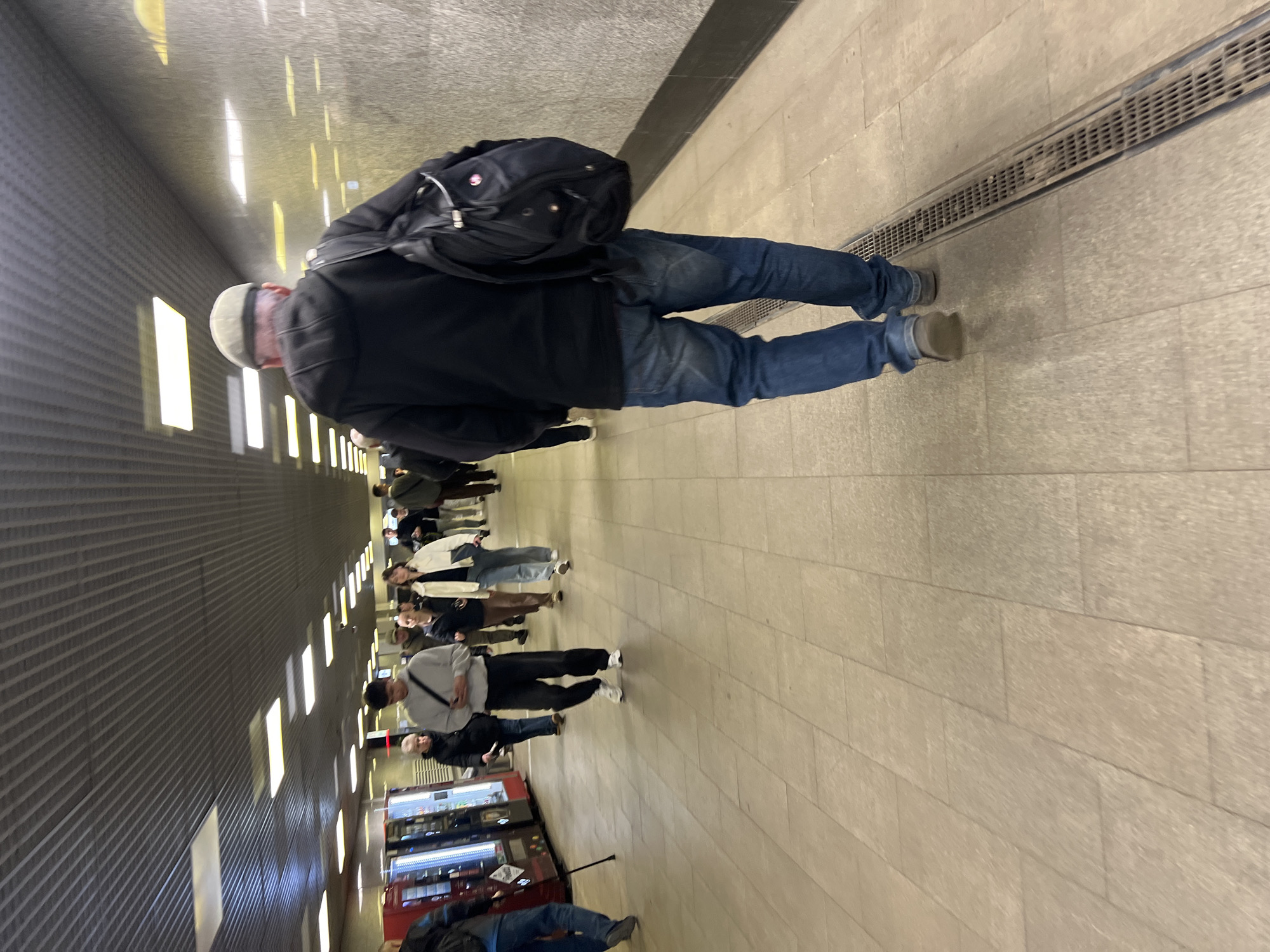 The image captures an indoor scene within a modern, well-lit hallway, likely a public transportation hub or a large building, potentially in Khimki, Russia. The perspective is from behind a man in the foreground, who is walking towards the end of the hall. He is wearing a black jacket, blue jeans, and a light-colored cap, carrying a black backpack.
A line of people extends further down the hallway, some appearing to be queuing or walking in the same direction, while others face partially towards the camera. They are dressed in casual attire, including jackets, sweaters, and jeans.
The hallway features a light-colored tiled floor with a long, narrow metal grate running parallel to the path on the right side. The left wall is dark gray with a textured, ridged pattern and is illuminated by evenly spaced rectangular light panels. Additional overhead lighting brightens the space. Along the right side of the hallway, a series of dark-colored vending machines or kiosks are visible. No specific text on these machines is clearly discernible. The scene depicts a functional public area, suggesting movement and waiting. The artificial lighting indicates an indoor environment where the time of day is not a factor.