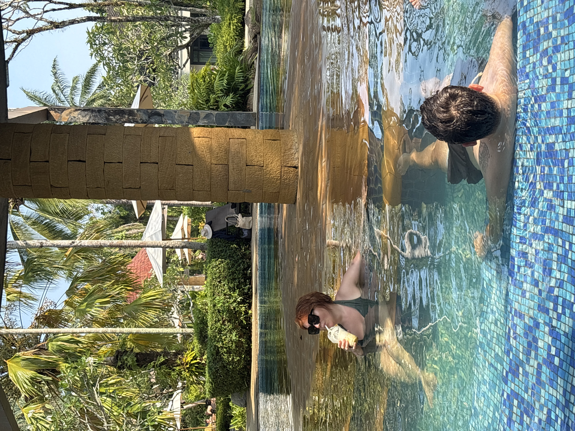 A woman in a green swimsuit and sunglasses is drinking from a cup in a pool, with a man partially submerged in the same pool. The pool is bordered by blue tiles on one side and a textured stone structure on the other. Lush green trees and vegetation surround the pool, and the sky is visible in the background. This location appears to be in Khok Kloi, Thailand.