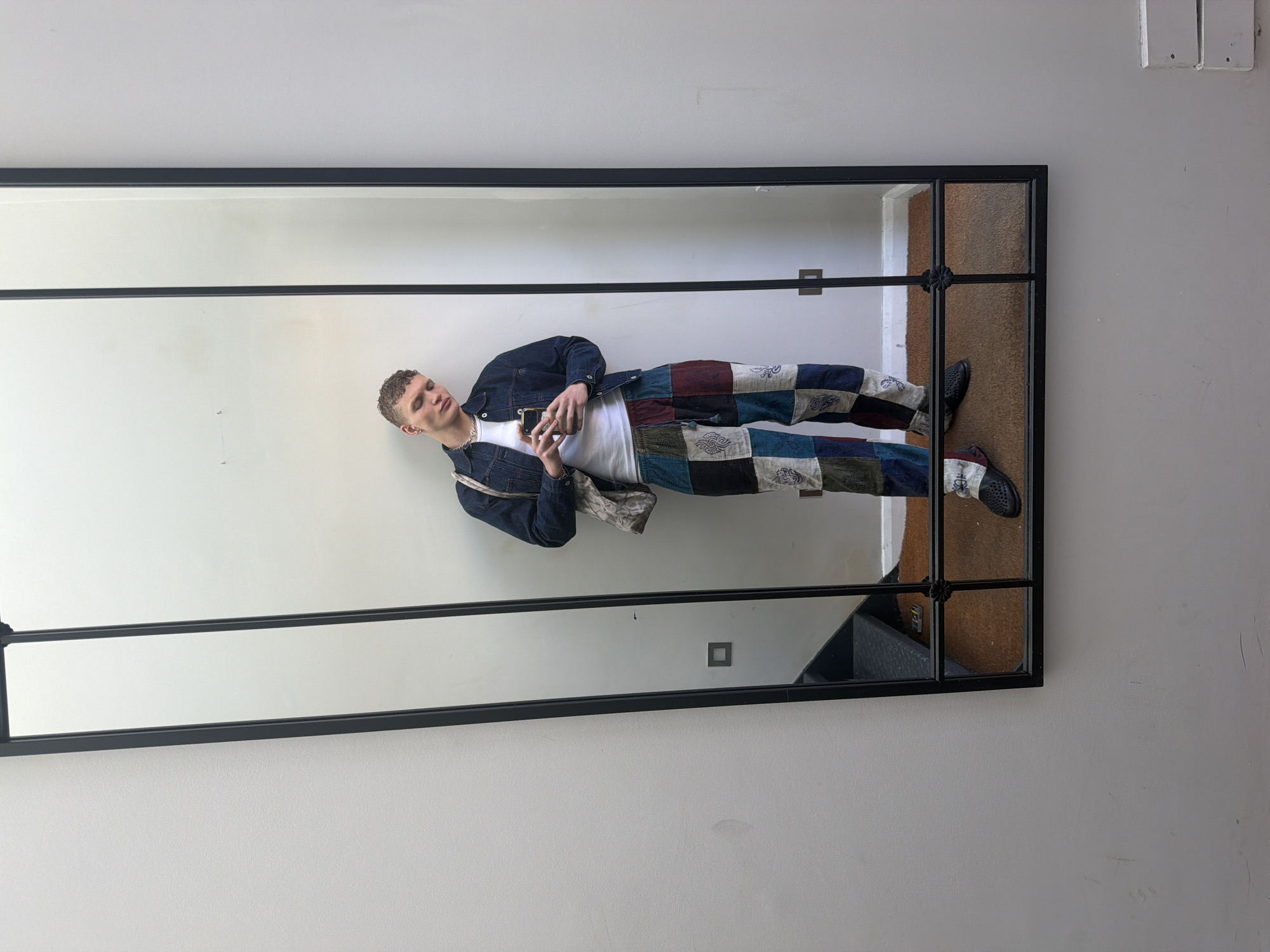 A young man wearing patchwork pants, a white t-shirt, and a denim jacket stands in front of a rectangular mirror with a black frame, holding a smartphone. He also wears a silver necklace. His reflection is visible in the mirror. The mirror is mounted on a white wall in what appears to be an interior space. The location is not specified, nor is the city of London, United Kingdom implied from the image. A brown rug and part of a staircase are visible in the reflection.