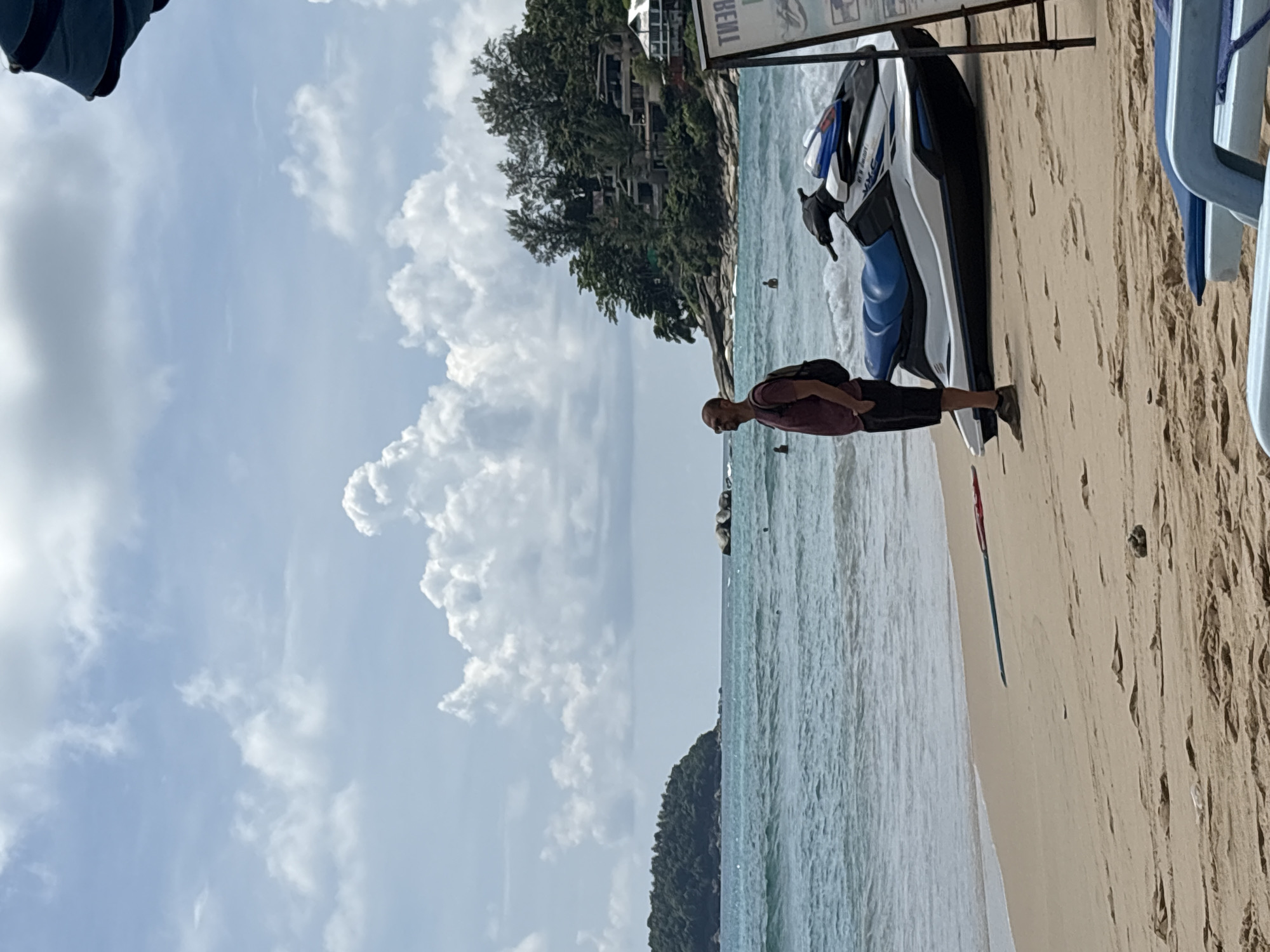 A man stands on a sandy beach in Karon, Thailand, looking out at the ocean. He is wearing a maroon t-shirt, black shorts, and brown shoes, and carries a black backpack. To his right, a white and blue jet ski is parked on the sand. The ocean is a vibrant turquoise, with gentle waves breaking on the shore. In the background, a lush green island rises from the sea, dotted with buildings. The sky is filled with scattered white clouds against a pale blue backdrop. The beach is relatively empty, with a few other people visible in the water. The overall atmosphere is serene and tropical.