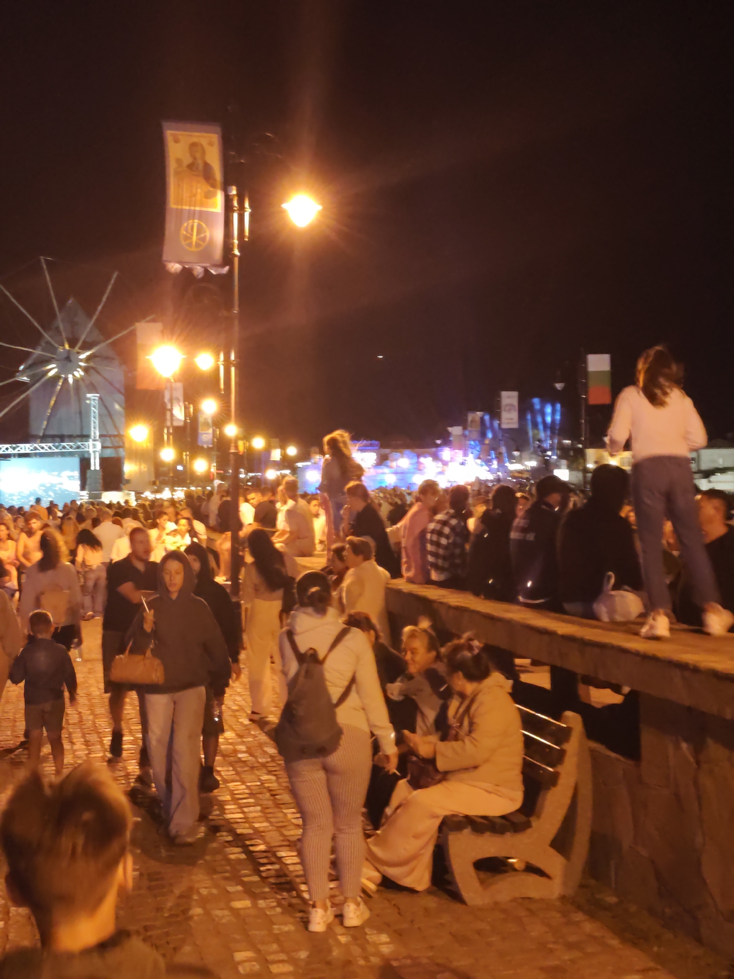 The image depicts a nighttime scene in the city of Nesebar, Bulgaria, likely during an event or festival. In the background, a Ferris wheel is visible, along with a stage setup and numerous people gathered in a street or plaza. Banners are hung from lampposts, one of which features a religious icon. Several streetlights illuminate the scene, casting a warm glow on the cobblestone pavement and the diverse crowd.
In the foreground and mid-ground, groups of people are walking and sitting. Some individuals are seated on a low wall, while others are gathered around a park bench. The people are dressed in casual attire, indicative of a public gathering. The atmosphere appears lively and social, with people engaged in conversation and observation. The overall impression is one of a community event taking place in a well-lit, pedestrian-friendly area of Nesebar.