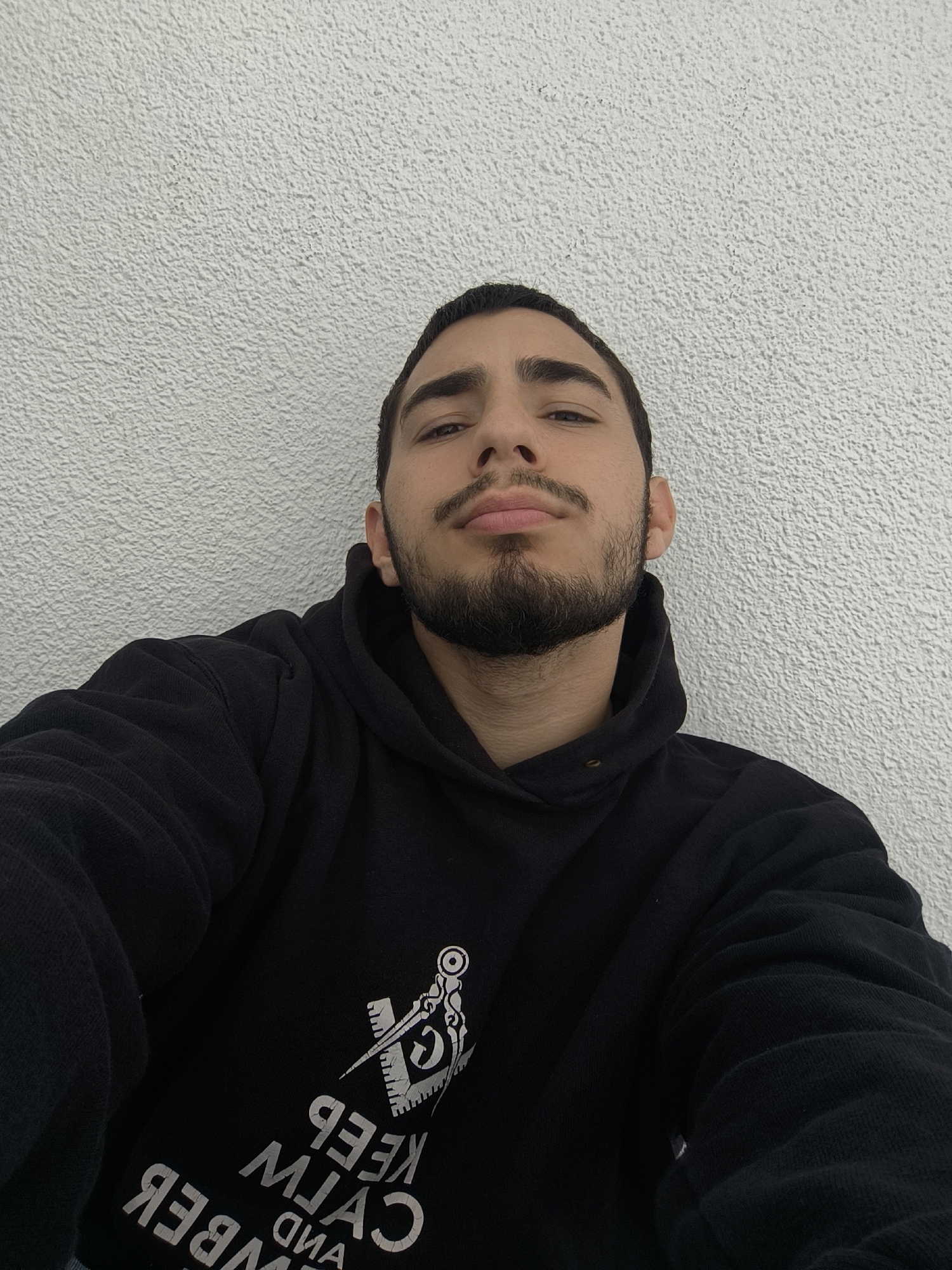 A medium shot of a man with a beard and mustache, facing the camera. He is wearing a black hoodie with white text and a symbol. The text appears to say "KEEP CALM AND REMEMBER". The background is a textured white wall. The location is not discernible from the image and cannot be confirmed as Schiltigheim, France.