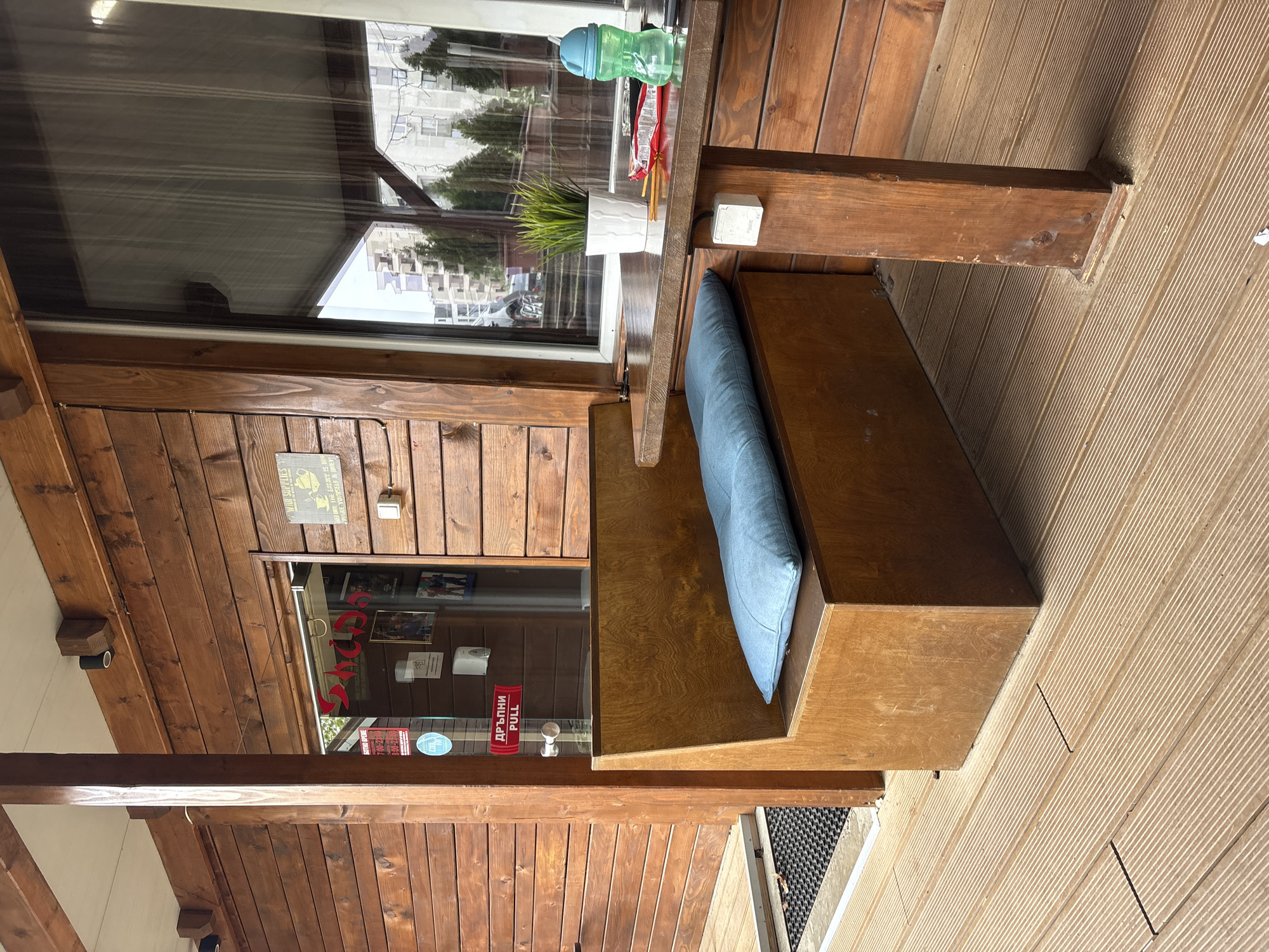 A partially outdoor seating area is photographed from a high angle. The location appears to be a cafe or restaurant in Sofia, Bulgaria. The predominant material in the photo is wood, finished with a brown stain. There are wooden planks on the left side and bottom, and a wooden bench with a blue cushion in the middle-right. Adjacent to the bench is a table, also made of wood, with a small plant, a water bottle, and a packet of something on it. Behind the table is a large window. To the left, a window shows a sign with a name beginning with 'S'. There are several other notices in the window, and a sign on the wooden wall above. The flooring is a grooved wooden material. The perspective is slightly distorted.