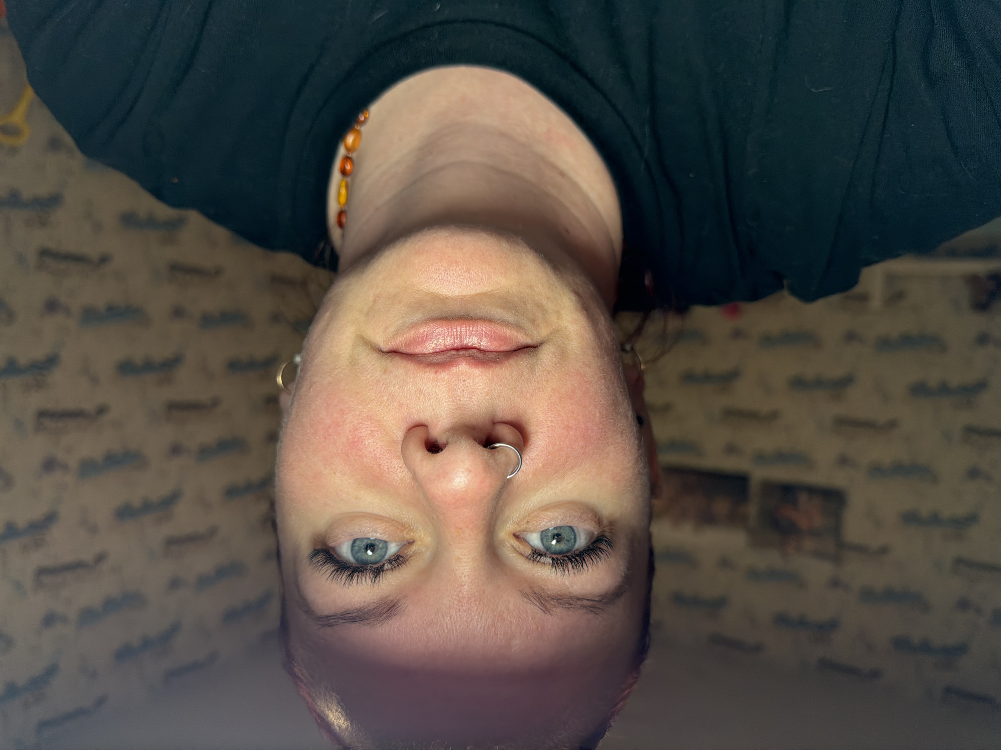 A close-up, inverted portrait of a light-skinned person with blue eyes is presented. The person has a septum piercing and earrings. They are wearing a dark-colored top and a necklace with orange-toned beads. The background appears to be a room with patterned wallpaper and blurry photographs. The location is presumed to be Chaikovsky, Russia.