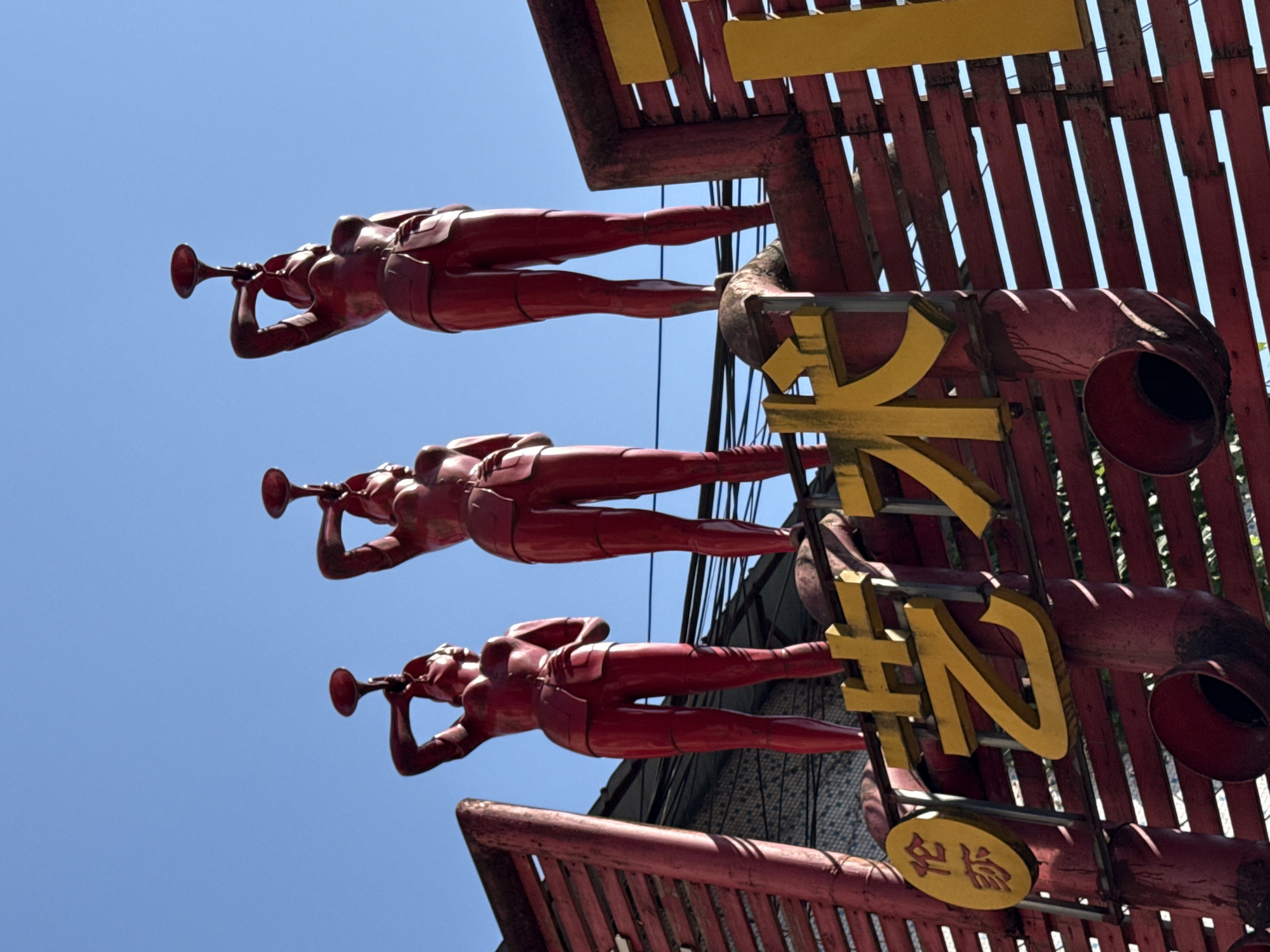 The photograph captures a low-angle view of a sculptural installation against a clear blue sky. Three identical, red-painted, stylized human figures are horizontally positioned in a vertical stack. Each figure is depicted in a supine pose, holding a trumpet to its mouth as if playing. The figures are mounted on a red, wooden structure adorned with large, yellow Chinese characters. Additionally, there's a small, round sign with more Chinese characters, also in yellow. The red wooden structure is made of parallel slats, creating a gridded appearance. This artwork is located in the Jiulongpo District of China.
