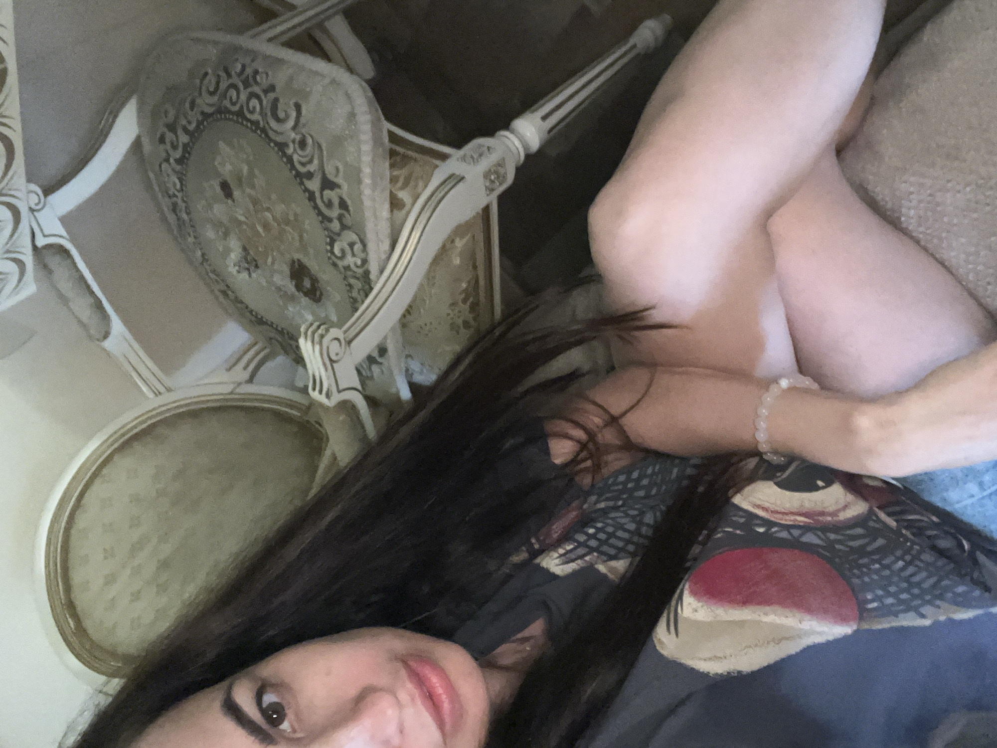 The image shows a person lying down, with their legs crossed. The person has long, dark hair and appears to be wearing a patterned shirt. Their face is partially visible, with one eye open and lips slightly parted. A beaded bracelet is visible on their wrist. In the background, there are two ornate chairs with light-colored upholstery and detailed carvings on the wood. The setting appears to be indoors, possibly a living room or dining room. The lighting suggests it might be daytime, but the exact time is unclear. There is no visible text in the image. The overall mood is relaxed and casual.