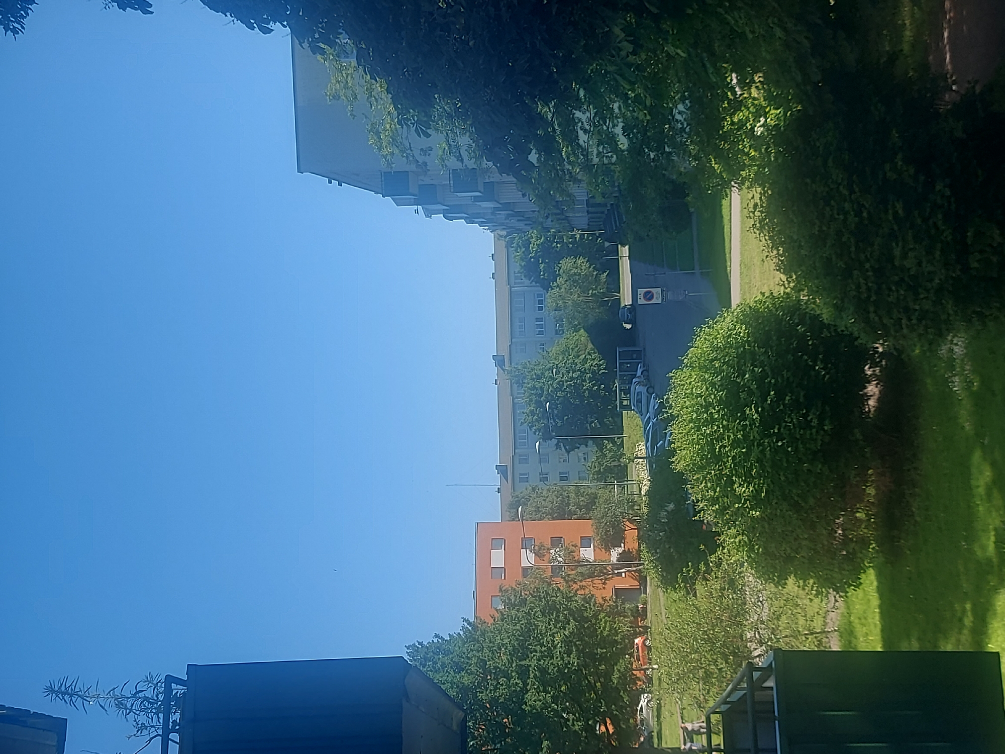 This is a landscape-oriented photo taken outdoors on a clear, sunny day. The image features a residential area with several multi-story buildings, some of which are orange and light gray. Lush green trees and bushes are abundant throughout the scene, casting shadows on the manicured lawns. In the foreground, parts of dark structures, possibly a building or a shed, are visible. A paved pathway or road runs between the buildings and the grassy areas, with parked cars and a traffic sign indicating a restricted area. The sky is a vibrant, cloudless blue. The scene appears peaceful, suggesting a typical day in a suburban or urban residential neighborhood. There is no visible text in the image. The orientation of the photo is rotated 90 degrees counterclockwise.