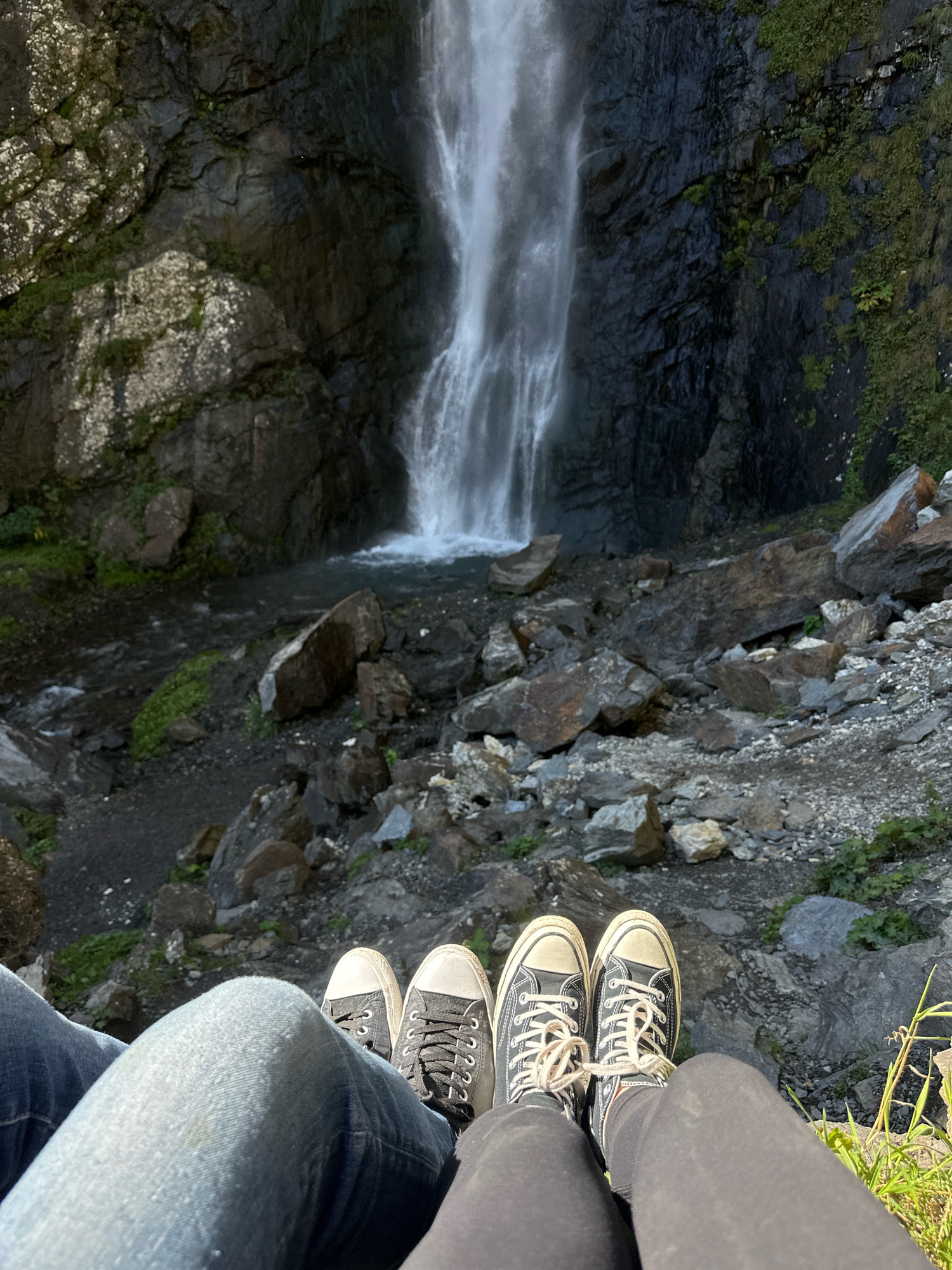 This is a photograph taken from a first-person perspective, showing a person's legs and feet in the foreground. They are wearing jeans and black Converse sneakers. Their legs are bent at the knees, and they are sitting with their feet extended towards a waterfall in the background. The waterfall is tall and cascades down a rocky cliff face covered in dark vegetation. The surrounding area is rocky and uneven, with large boulders scattered around the base of the waterfall and in the pool of water at its foot. The scene appears to be a natural outdoor setting, likely a tourist spot given the presence of a paved path visible on the left side of the frame. The location is identified as Tsdo, Georgia.