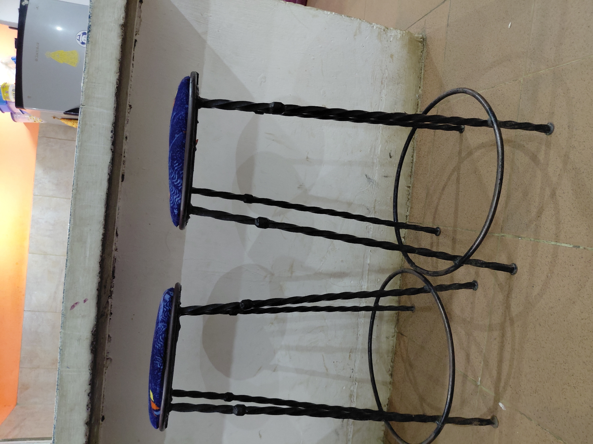 The image displays an indoor scene, likely a kitchen or bar area, featuring two black wrought iron bar stools positioned next to a counter. The stools have intricately twisted metal legs and supports, with circular footrests near their bases. Each stool is topped with a round, blue upholstered seat featuring a light-colored, swirling or abstract pattern.
To the left, a light-colored counter with a rough, unfinished edge spans horizontally. Behind the stools, a plain white or off-white wall shows some minor marks and discoloration. The floor, covering the lower and right portions of the frame, is composed of light brown or beige square tiles with visible grout lines.
In the background, on the far left side, a silver-colored appliance, identified by the visible text "Samsung," is partially seen above the counter. On top of this appliance, another item shows the text "ELECTROLUX." A distinctive yellow sticker, depicting a stylized figure that could be interpreted as a cartoon character or a religious icon, is affixed to the refrigerator door. Various bottles and containers are also visible on the counter and around the appliance.
No people are present, and no specific activity is taking place, making it a static interior view. The lighting suggests daytime conditions. Given the provided context, the scene is set in Santa Ana, Venezuela, depicting a functional, possibly domestic or small commercial space.