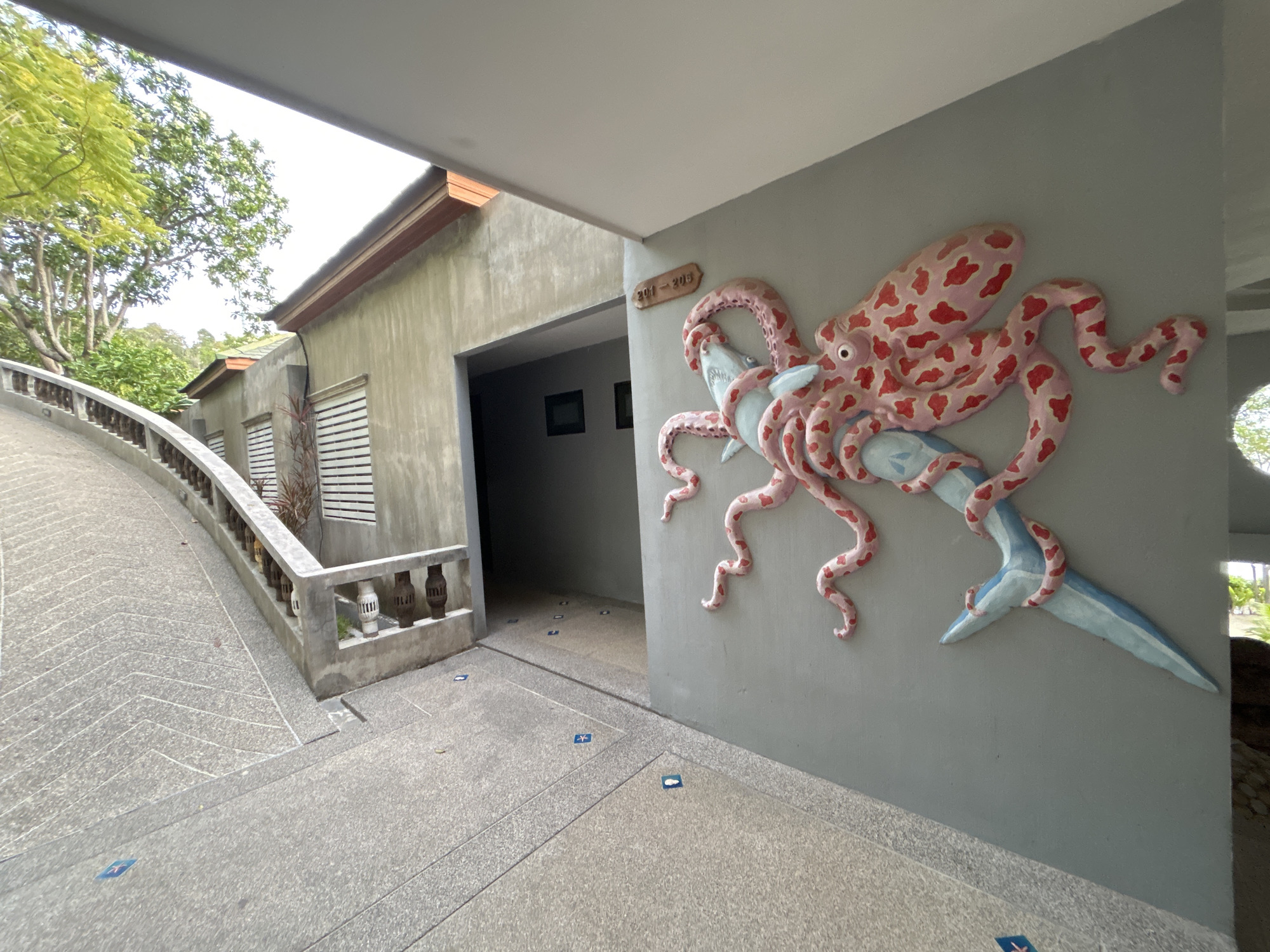 The image shows a building entrance in ตำบลเกาะพะงัน, Thailand. A concrete ramp leads up to a walkway, and a building with a gray exterior is visible. A sign reading "201-205" is above the entrance to a hallway. A large, three-dimensional sculpture of an octopus attacking a fish is on the wall adjacent to the entrance. The ground is paved with gray stone.