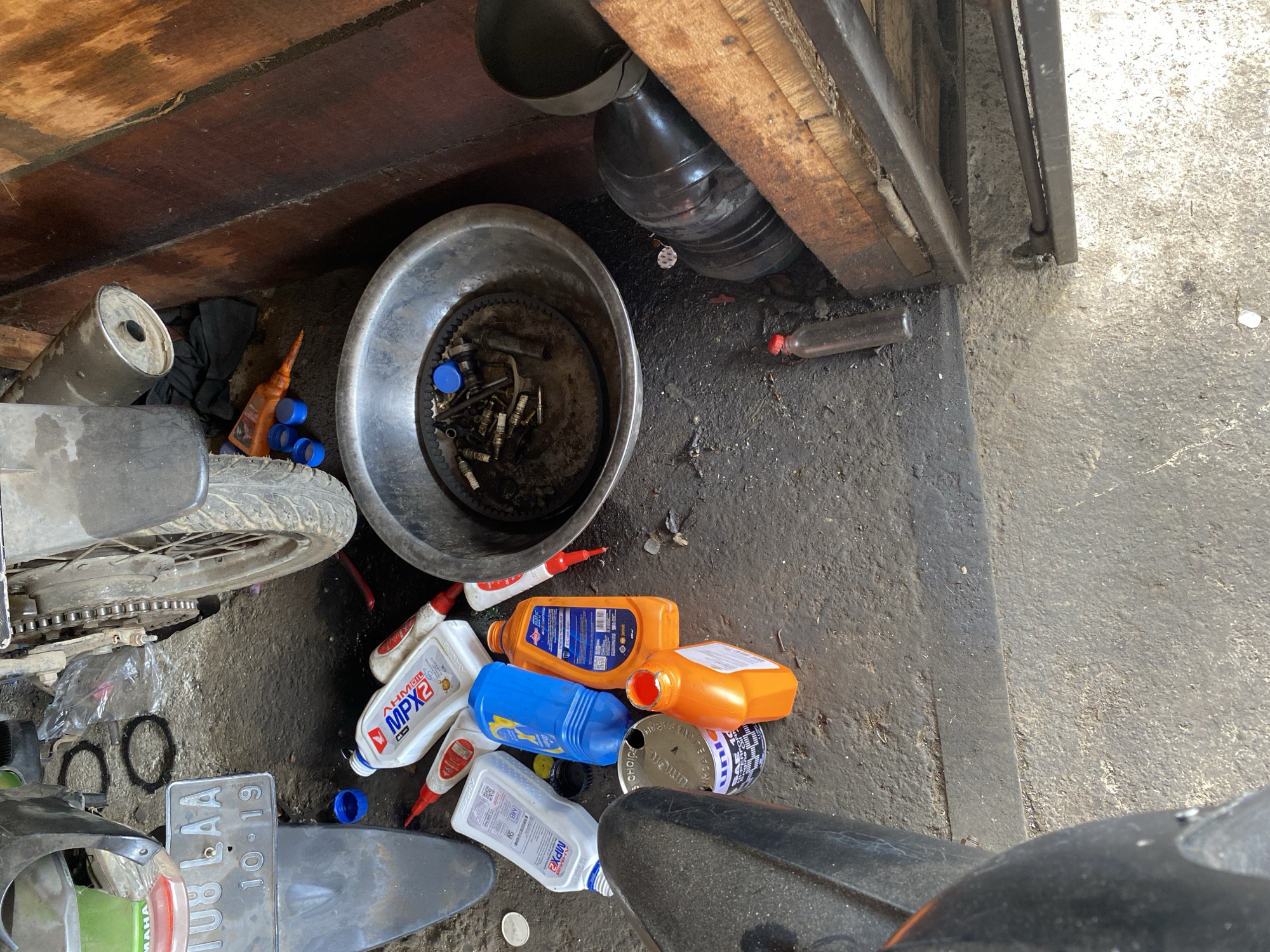 In Bayu, Indonesia, an outdoor motorcycle repair area is depicted with various items scattered on a rough, gray concrete ground. A partially visible motorcycle is present, with its rear tire, chain, and part of the exhaust visible. Several containers, including an orange plastic bottle with a red cap, a blue plastic bottle, and white bottles labeled "MPX2," are on the ground, suggesting a motor oil change or repair. A metal basin filled with motorcycle parts, including spark plugs and a belt, sits in the center of the frame. A dark gray funnel and a large black plastic container are to the right. The background features a wooden structure with visible grain and stains. The scene appears to be a disorganized workspace.