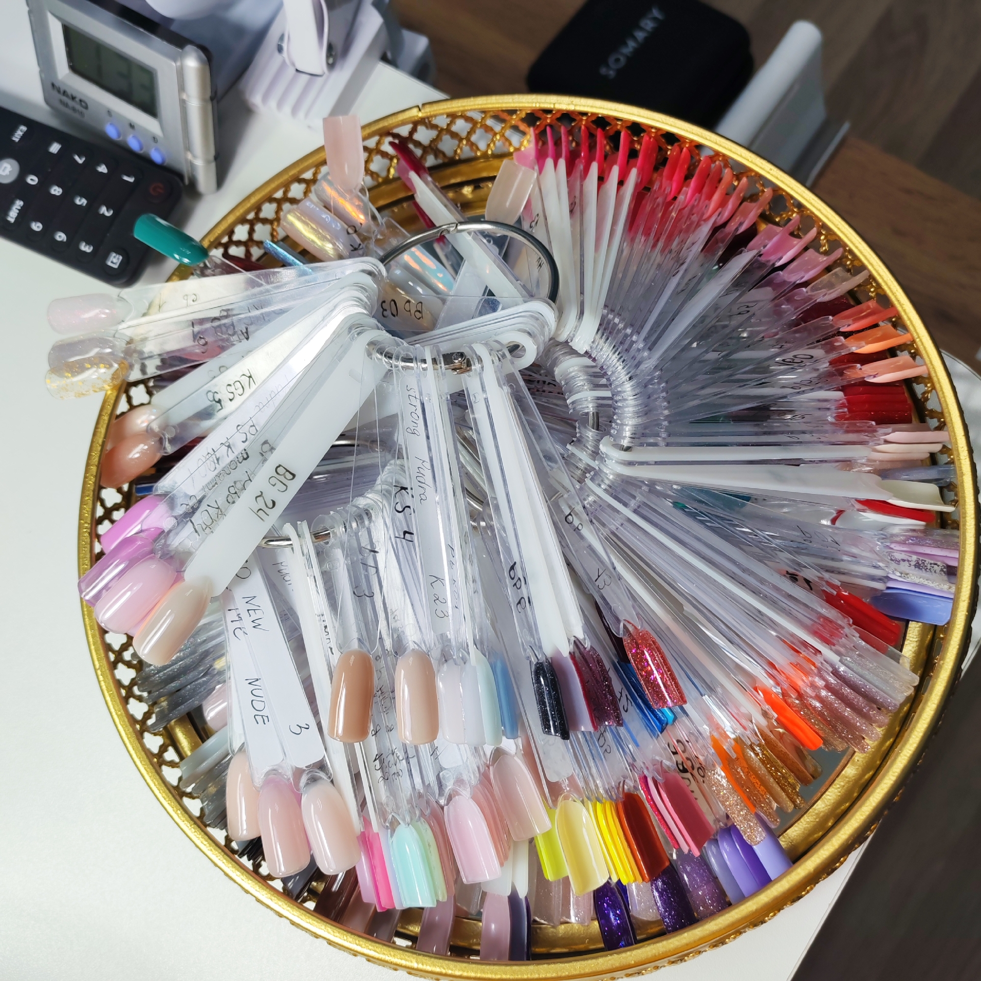The image shows a golden tray filled with nail polish color samples attached to plastic sticks and metal rings. The sticks have handwritten labels on them with brand names and color codes, like "BG 24," "KS 4," and "NUDE 3." A remote control and digital clock are in the background, along with nail care equipment. The label "SOMARY" is also visible, indicating a brand or business name. The scene depicts a collection of nail polish samples, likely in a salon or nail care setting. It is not possible to identify the location in Chaikovsky, Russia, based on the visual information alone.