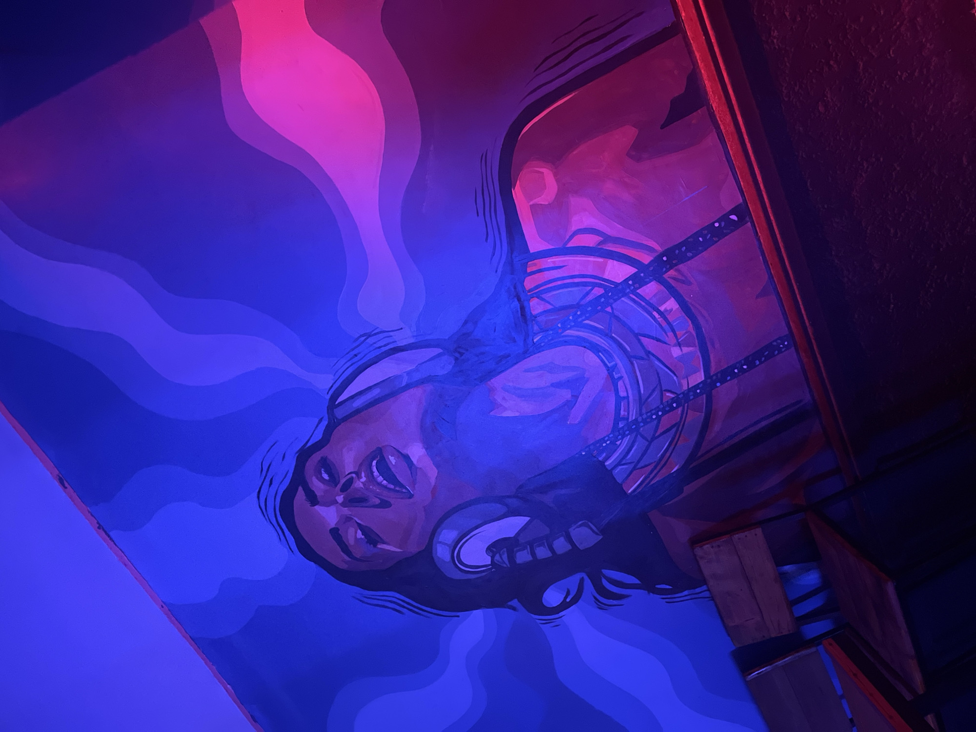 A mural of a woman wearing headphones is painted on a wall in Howayi, Nigeria. The mural is illuminated by blue and pink lights, creating a vibrant and atmospheric scene. The woman's eyes are closed, and her mouth is open, suggesting she is deeply engrossed in the music. She wears a necklace with intricate beadwork. In the foreground, several wooden chairs are visible, indicating this is a public space, possibly a bar or a lounge. The background of the mural features abstract wavy patterns in varying shades of blue and purple, transitioning to pink on the right side. The lighting casts strong shadows and highlights, enhancing the artistic effect of the mural.