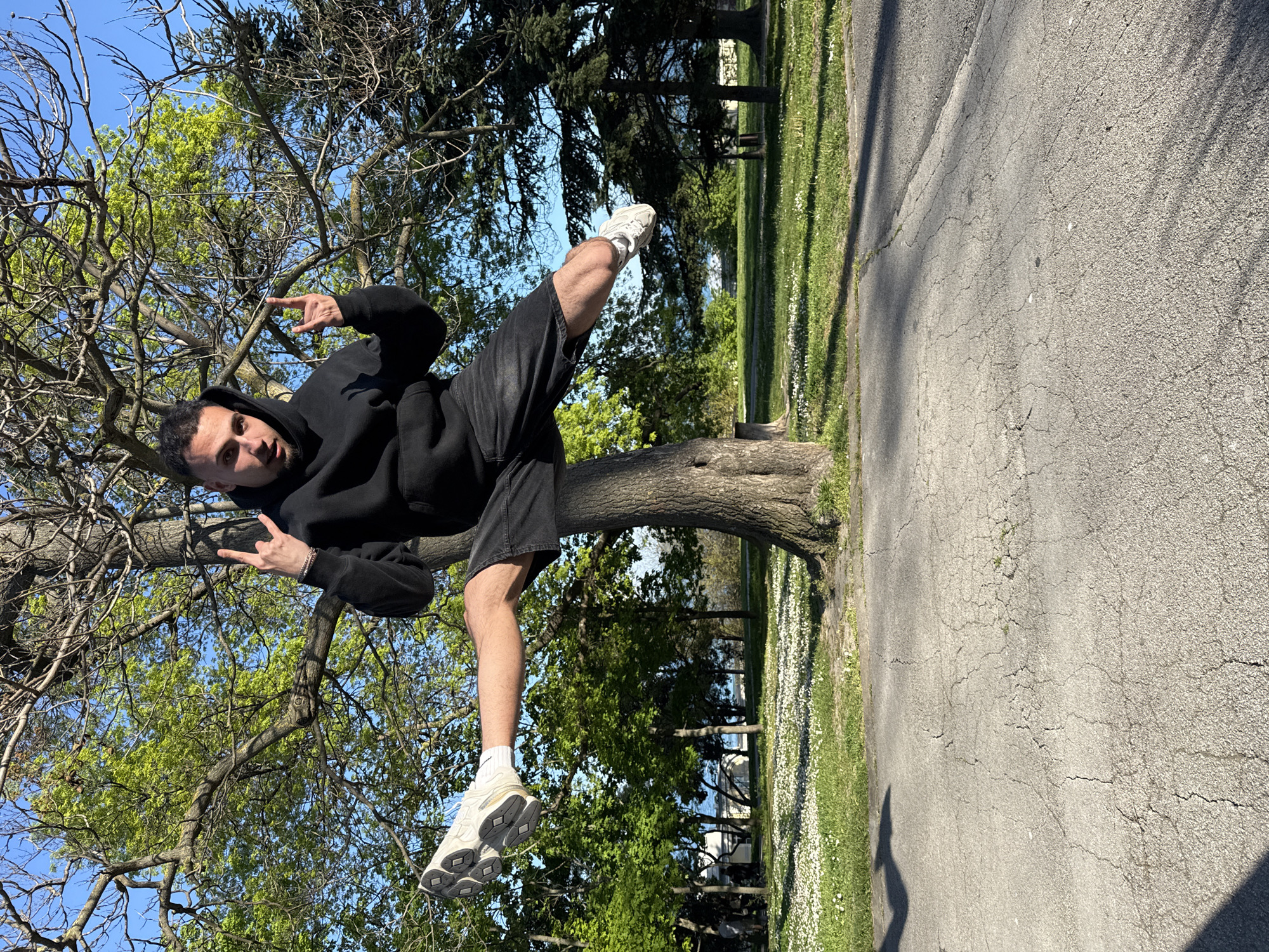 A man in a black hoodie and black shorts is sitting in a tree in a park. He is wearing white sneakers and holding up two fingers in a "rock on" gesture with both hands. The park has green grass and trees. The background includes a paved road. The location appears to be a public park with trees, grass, and a paved pathway. The location might be in a park in Varna, Bulgaria.