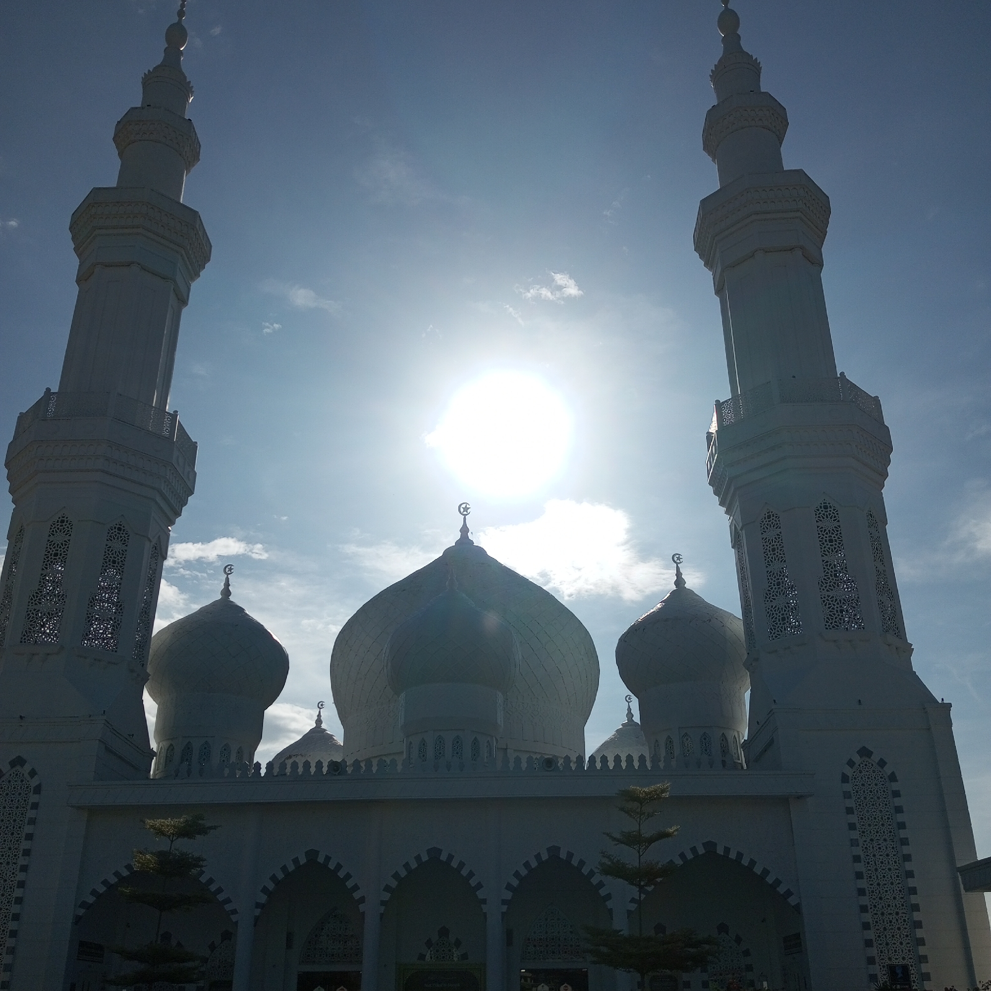 The image depicts Baitul Muttaqin Mosque in Meunasahaluedrienlhoksukon Barat, Indonesia, under a bright, sunny sky. The mosque is white, featuring two tall minarets with decorative latticework, several domes of varying sizes topped with crescent moons, and arched entrances. The sun is positioned directly above the central dome. Two small trees are in front of the mosque.