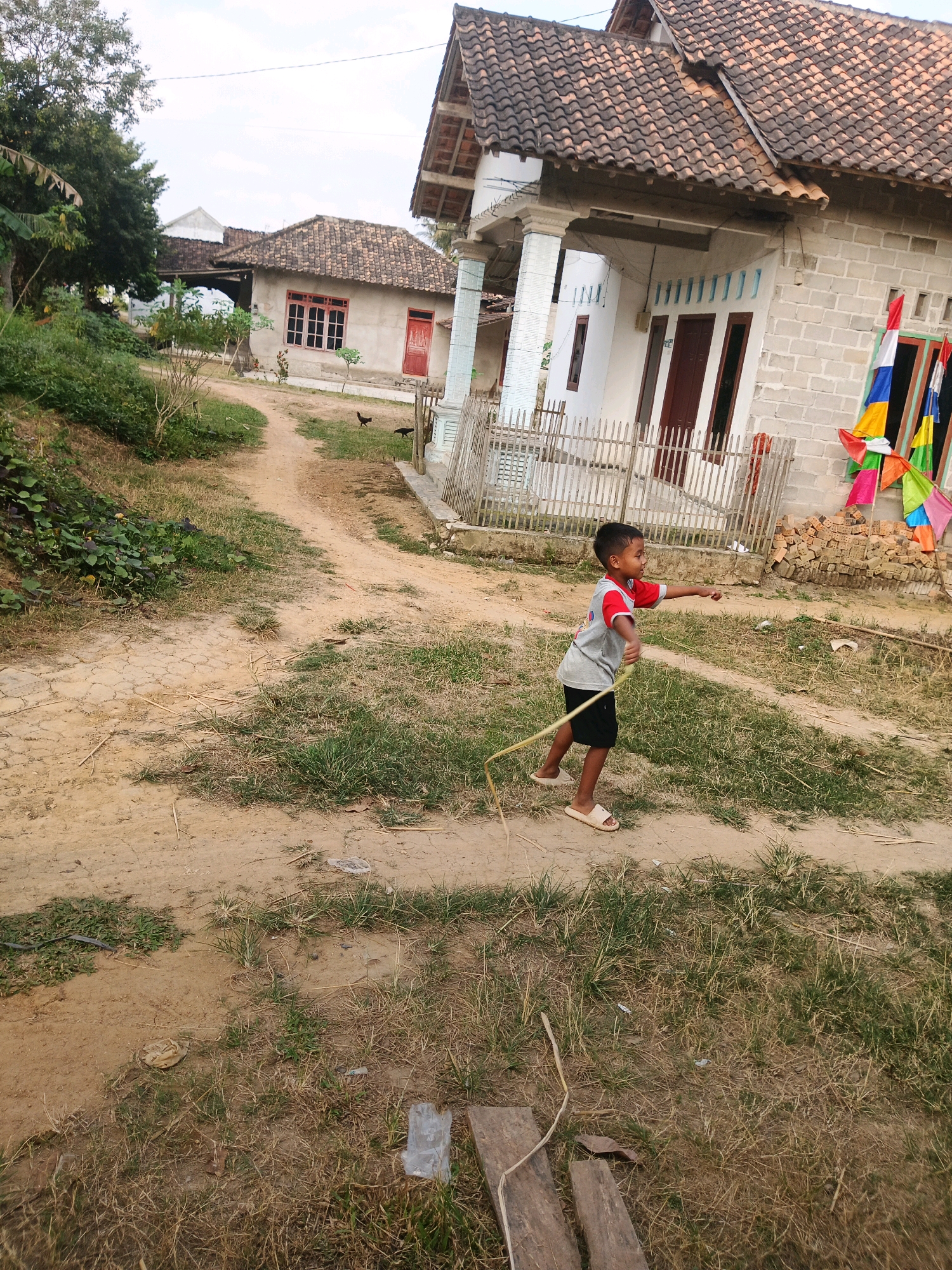 A young boy, wearing a gray t-shirt with red sleeves and black shorts, stands on a dirt path in front of a house in an unspecified village in Indonesia. He holds a yellow rope in his right hand, which extends behind him. His left arm is extended forward, and he appears to be in motion, possibly playing or skipping. The house behind him has a white brick facade, a brown tiled roof, and several windows with brown frames. Colorful flags are attached to the front of the house. In the background, another house with a similar tiled roof is visible, along with some greenery and trees. Two chickens are present on the path near the houses. The ground is a mix of dirt and dry grass.