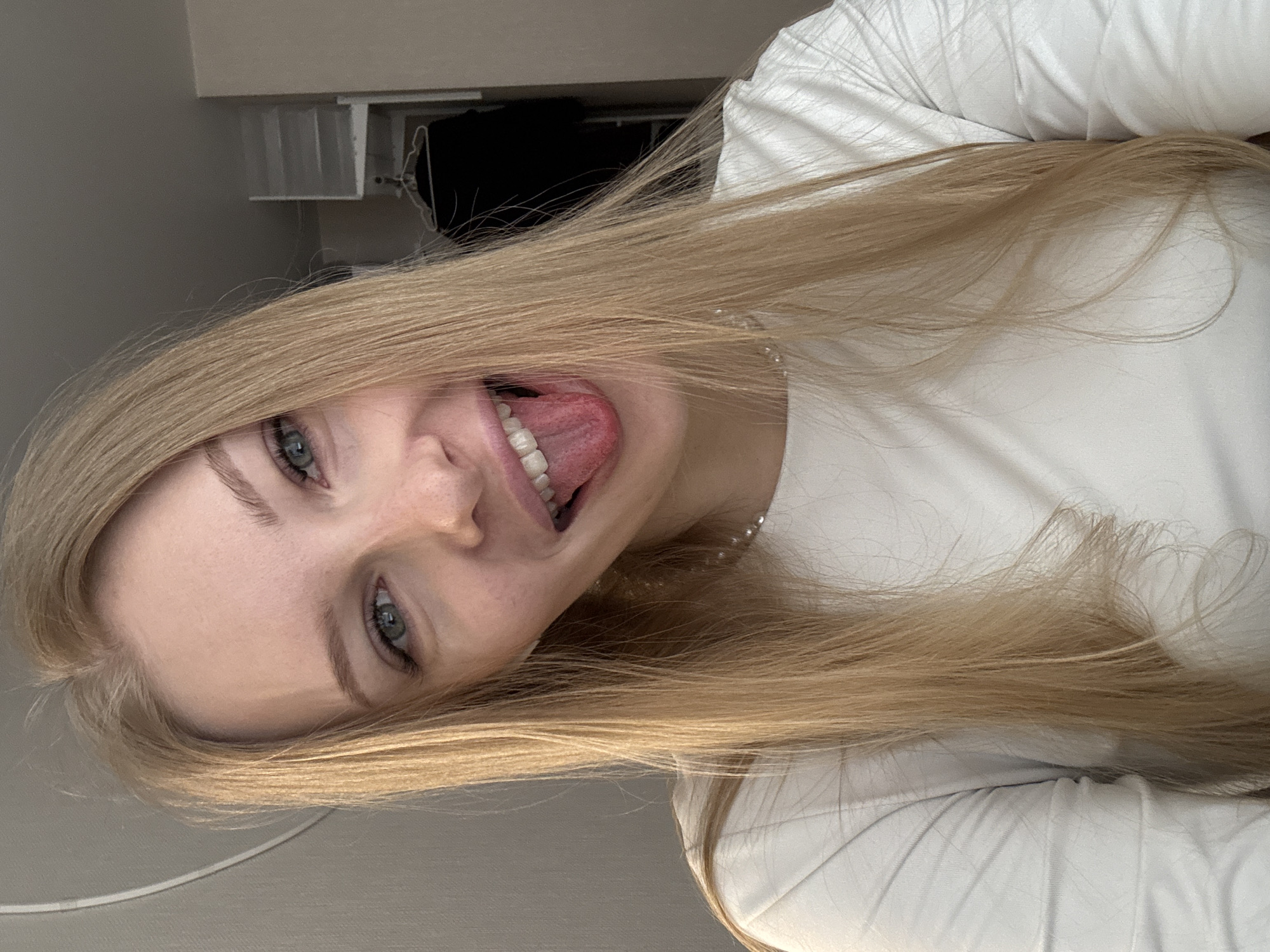 A young woman with long, straight blonde hair and fair skin is the central subject of the image. Her light blue-grey eyes are looking directly at the camera, and she is captured in a playful moment, sticking her tongue out with a cheerful expression. She wears a simple white long-sleeved top, and a pearl necklace is subtly visible around her neck. Her blonde hair cascades around her face and extends across the lower portion of the frame.
The setting appears to be indoors, likely a bedroom, indicated by the presence of what looks like white bedding or a pillow towards the right side of the image. The background features a light-colored wall, and in the upper portion, indistinct domestic elements such as parts of a window frame, a shelf, or furniture, along with a dark area that might be a curtain or hanging clothes, are visible. The lighting is soft and even, suggesting indoor ambient light, possibly during the daytime. No specific time of day or weather conditions are discernible from the interior shot. The woman's emotion is clearly playful and lighthearted. There is no visible text in the image. The location context provided is Perm, Russia.