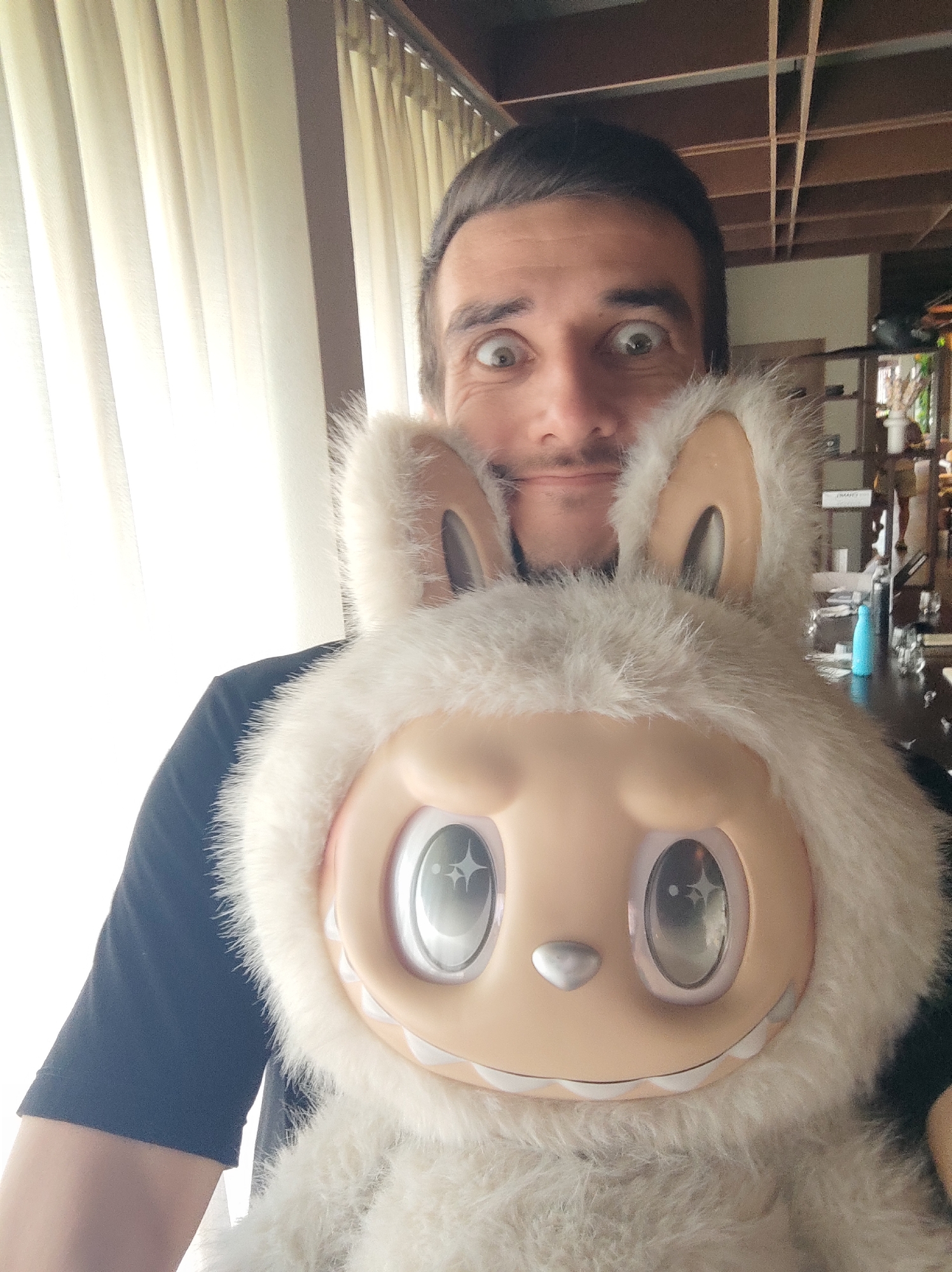 A man takes a selfie indoors while holding a large, fluffy, rabbit-like plush toy. The man, positioned in the upper center of the frame, has dark hair and a light beard. His eyes are wide open in a playful or surprised expression, complemented by a slight smirk. He is wearing a dark, short-sleeved t-shirt.
The oversized plush toy dominates the lower and central part of the image. It features a creamy, cartoonish face with large, shiny, star-speckled eyes and a small silver nose. A jagged line of white, tooth-like details forms its grin. The toy is covered in shaggy white fur, giving it a soft, ethereal appearance, with light brown inner ears protruding from the fur.
The setting appears to be a well-lit indoor space during the daytime. To the man's left, a bright window with light-colored vertical blinds or curtains is visible, allowing ample light into the room. To his right, the background is darker, revealing a wooden slatted ceiling structure and various shelves or furniture. A dark counter or table holds a light blue bottle and some glasses, suggesting a casual living area, cafe, or bar. The image captures a lighthearted, humorous moment. No text is visible. This scene is contextualized as taking place in North Kuta, Indonesia.