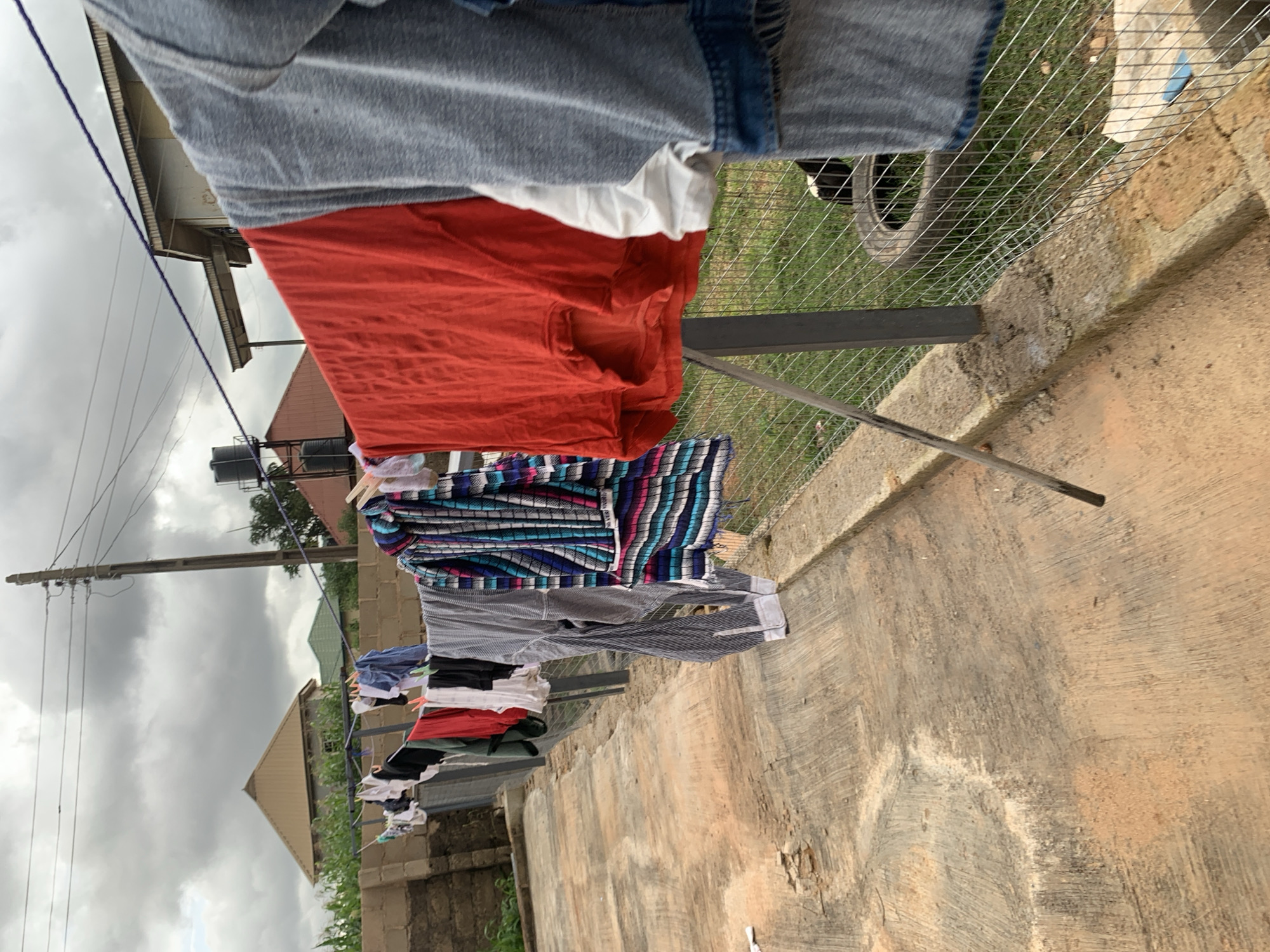 An outdoor scene depicts laundry hanging on a clothesline. A variety of garments, including shirts, pants, and what appears to be a dress or skirt with horizontal stripes in blue, pink, and white, are suspended on the line, secured by clothespins. The clothesline is supported by metal poles. In the foreground, a concrete pathway with visible wear and tear leads away from the viewer. Behind the clothesline, a wire fence encloses a grassy area where a tire and a black and white chicken are visible. Beyond the fence, buildings with various roof designs and a water tank can be seen, set against a backdrop of a cloudy sky. Power lines and a utility pole are also present in the upper portion of the image.