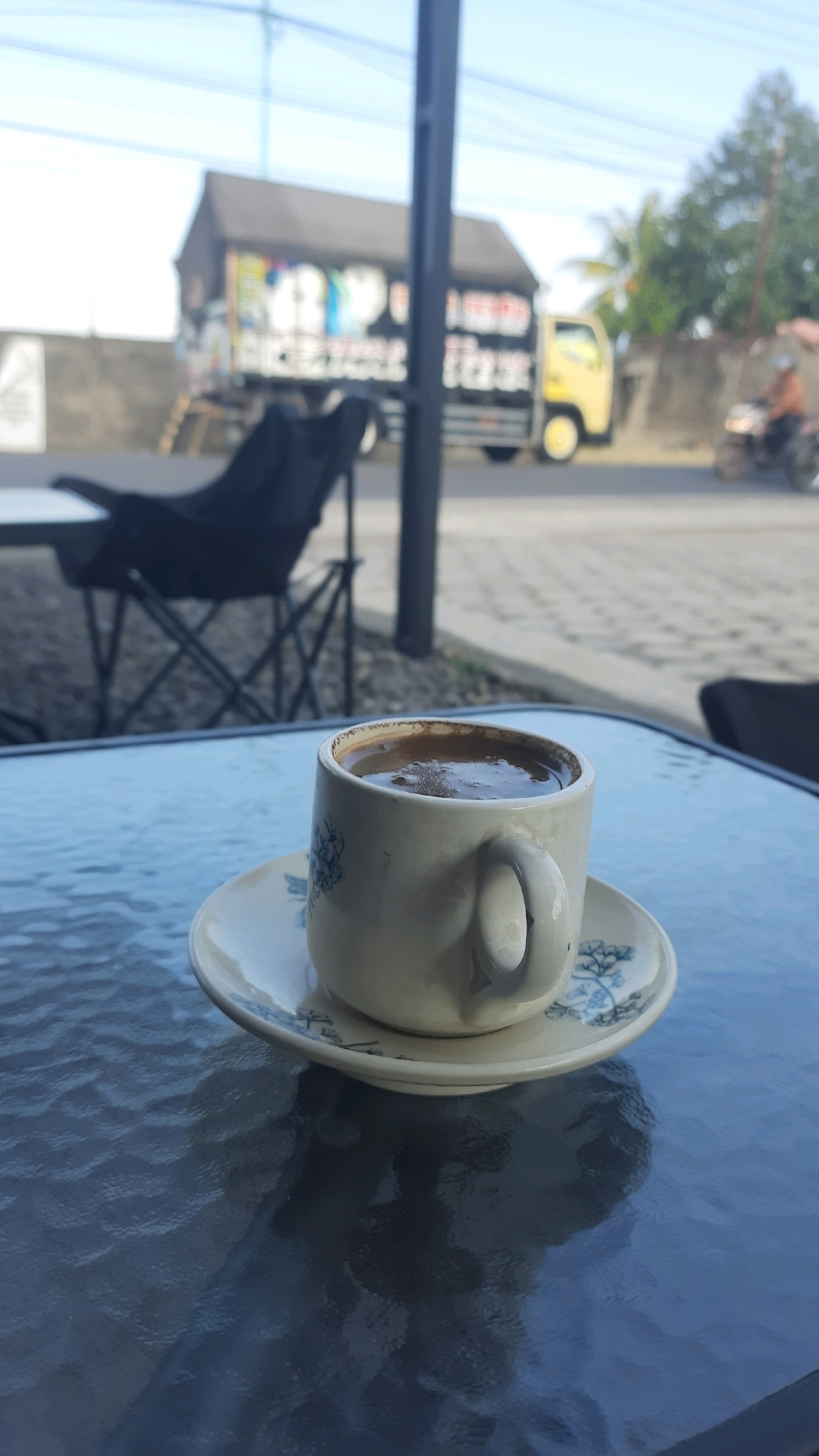A close-up, eye-level, outdoor shot features a cup of coffee on a textured, dark blue glass table. The cup is off-white with blue floral patterns and sits on a matching saucer. The coffee is dark and frothy. In the background, out of focus, is a street scene. A black folding chair is visible to the left, and a yellow truck with graphics on its side is to the right. A person on a motorcycle is further in the distance. The sky is bright blue. The location is Jajag, Indonesia.
