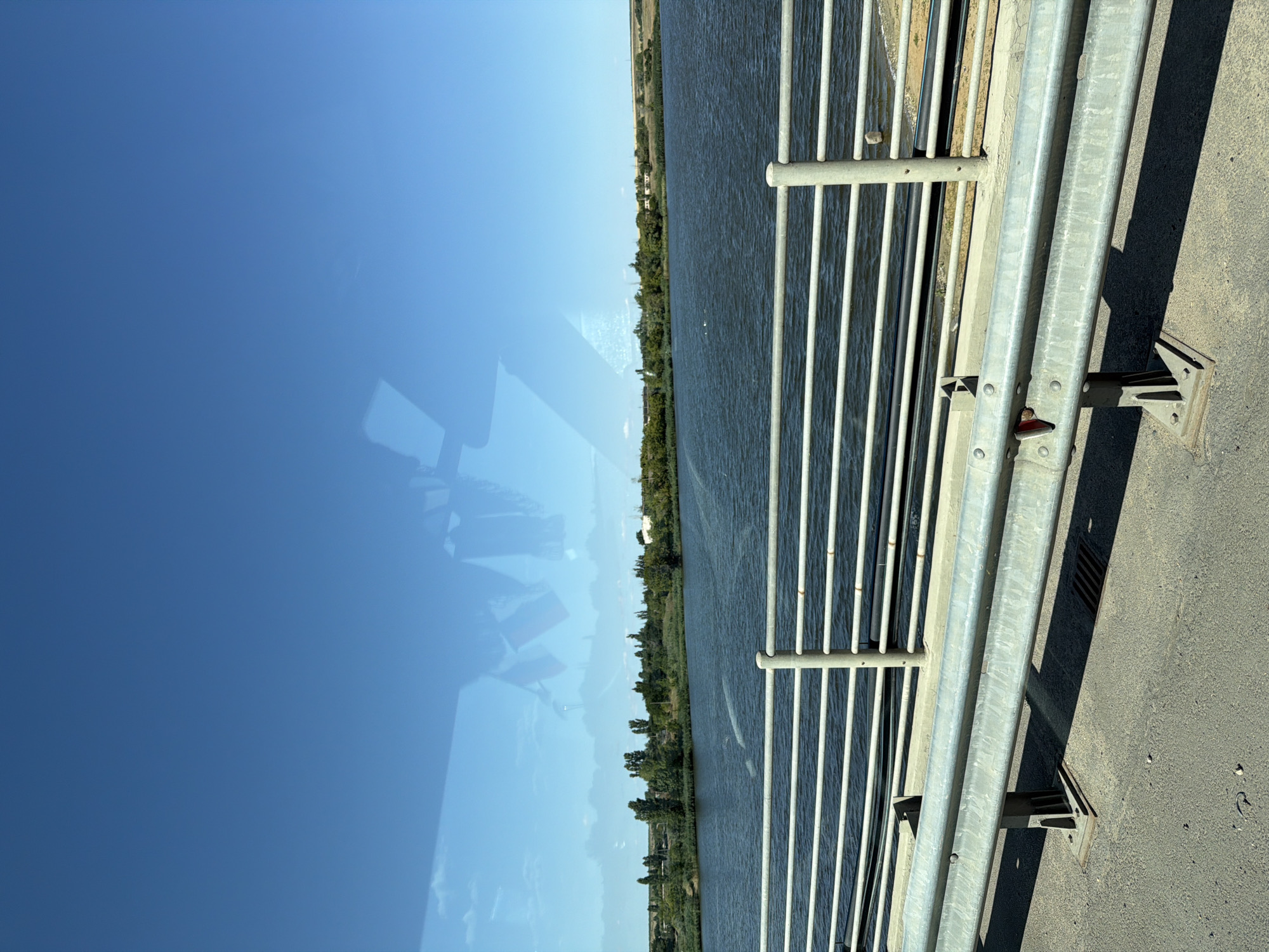 A wide shot captures a view from a bridge overlooking a body of water and a distant shoreline lined with trees and vegetation under a clear blue sky. In the foreground, a metal guardrail with horizontal bars runs along the side of the bridge, with a concrete barrier below it. The pavement of the bridge is visible, showing a drain grate. The water is dark and reflects the sky. The shoreline is densely covered with trees and shrubs, with a few buildings barely visible in the distance. The image appears to be taken during daylight hours, with bright sunlight.