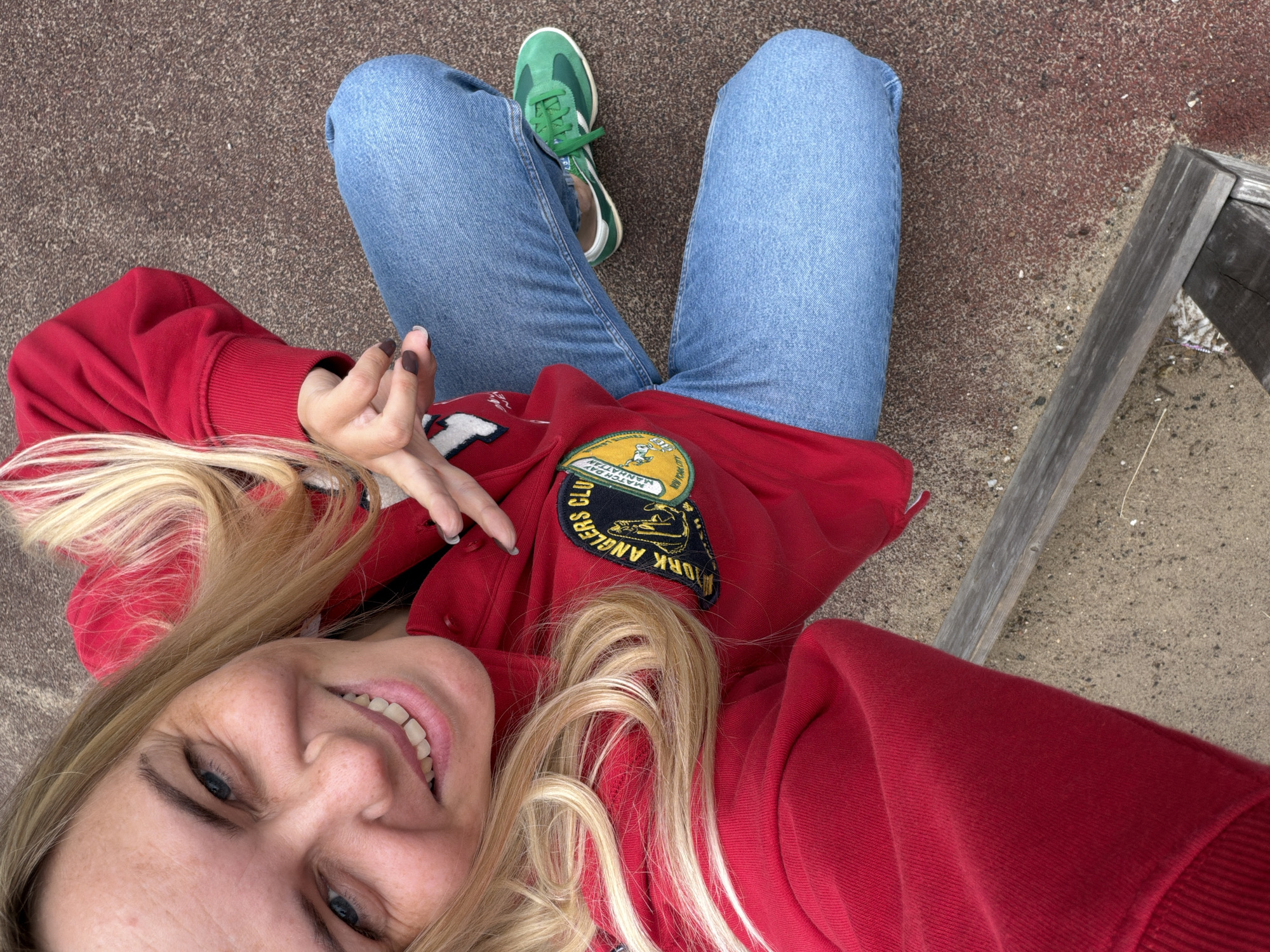 In an outdoor setting on a surface that appears to be a mixture of sand and red asphalt, a person is captured from a high-angle perspective. The individual, who appears to be female, has blonde hair and is wearing a red hooded sweatshirt with a patch on the left chest. The patch has text and graphics on it, but is partially obscured. The person is also wearing blue jeans and bright green athletic shoes. They are seated with their legs bent and crossed, and their left hand is raised, forming a gesture. The background shows a wooden beam or bench in the lower portion of the image. The overall scene suggests a casual outdoor activity. It is not possible to determine the specific location within the city of Votkinsk, Russia from the visual information provided.