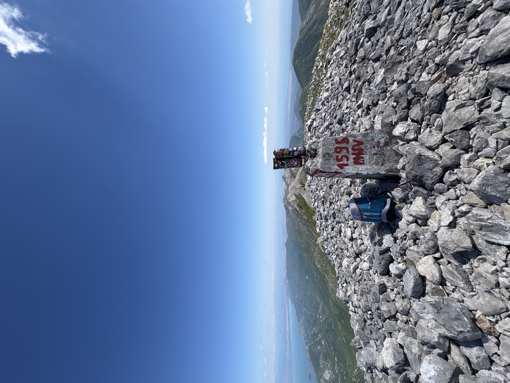 The image shows a mountain peak in Mali Mikulići, Montenegro. A concrete pillar, painted with "1595 MNV" in red, stands amongst a rocky terrain. The pillar also has several stickers attached to its top. A blue backpack rests against the pillar. In the background, the sky is clear blue, with a distant view of mountains and a body of water.