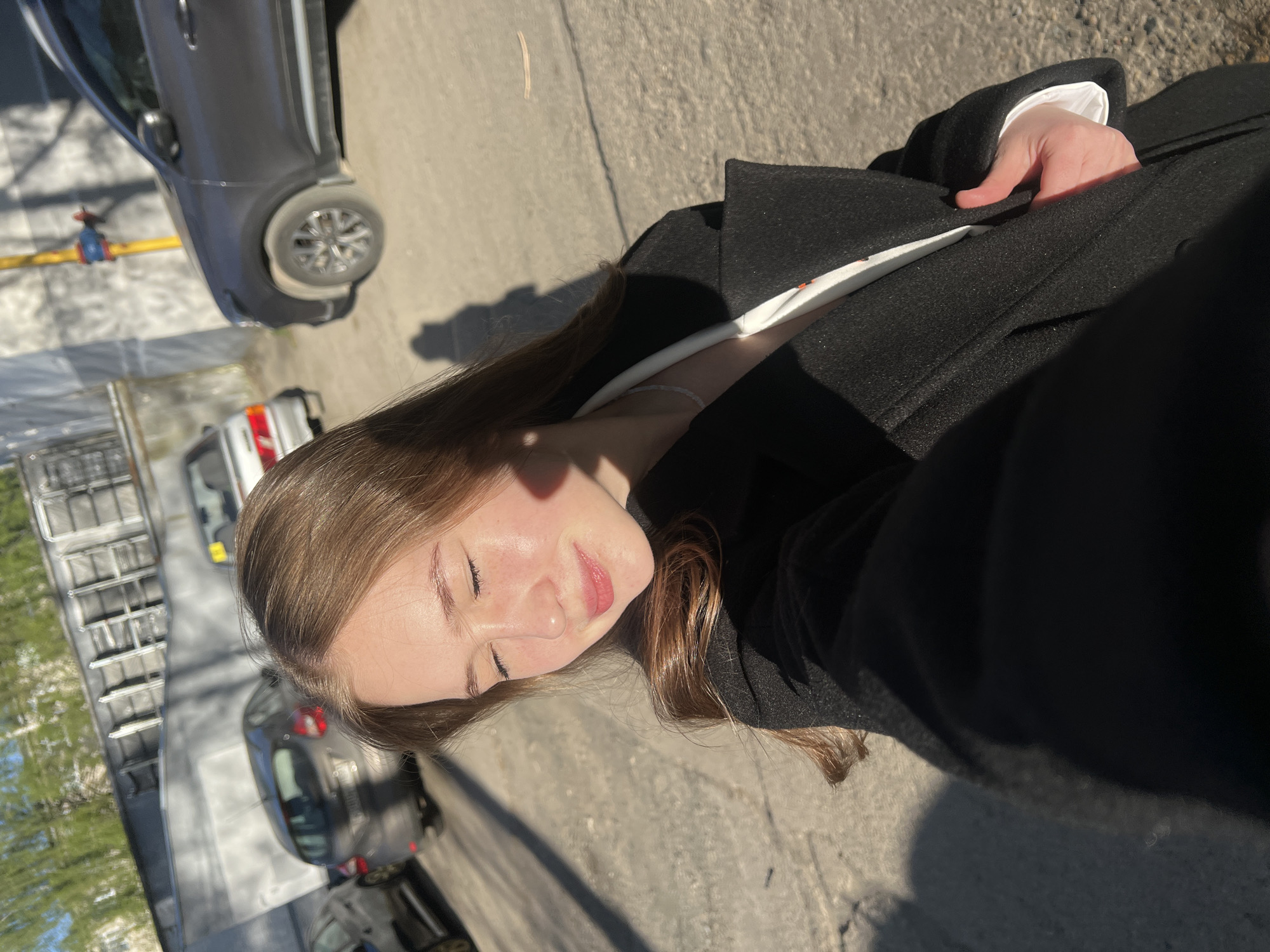 A young woman with light brown hair and closed eyes is taking a selfie outdoors. She is wearing a black coat with a white collar visible. The photo appears to be taken on a street in Chaikovsky, Russia. The background includes parked cars, buildings with balconies, and trees. The light suggests it is a sunny day.