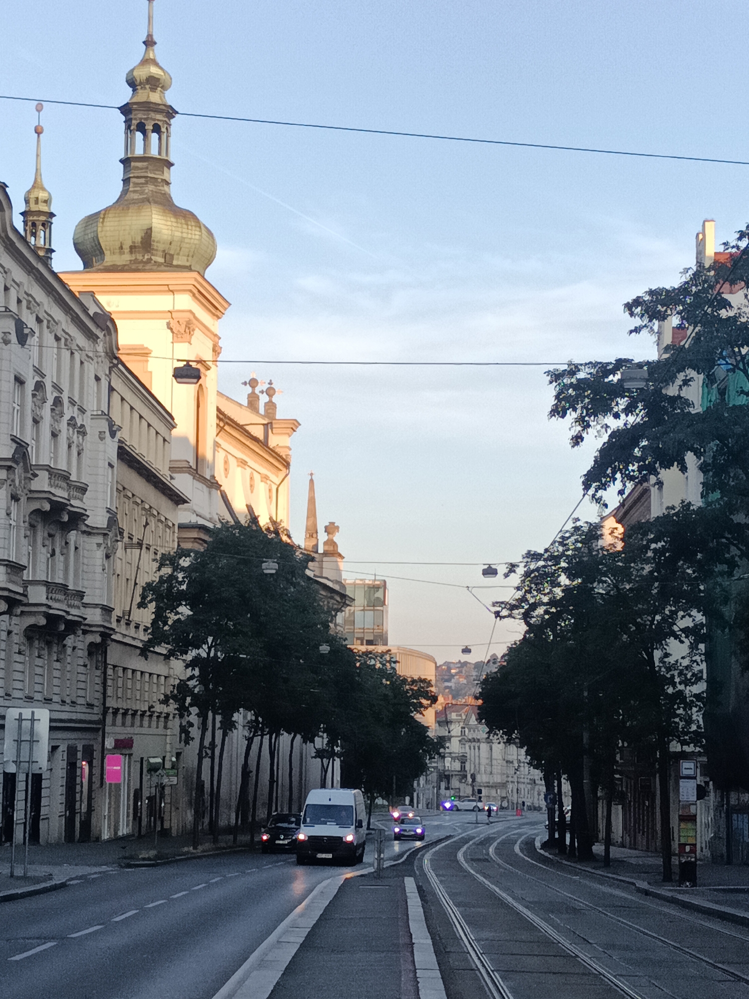 The image depicts a street scene in Prague, Czechia, featuring a tram line running through a European city. In the foreground, a white van is visible on the road, followed by a dark-colored car. To the left, the ornate facades of historic buildings line the street, with a prominent church tower topped by a golden dome dominating the skyline. Trees with dark foliage are strategically placed along the sidewalk, casting shadows. The tram tracks run parallel to the road, curving towards the background where more buildings and a hint of a hilly landscape are visible. The sky is a pale blue with wispy clouds, indicating either early morning or late afternoon light. Overhead wires, typical of tram infrastructure, crisscross the scene. The overall impression is one of a functioning urban environment with a blend of historical architecture and modern transportation.