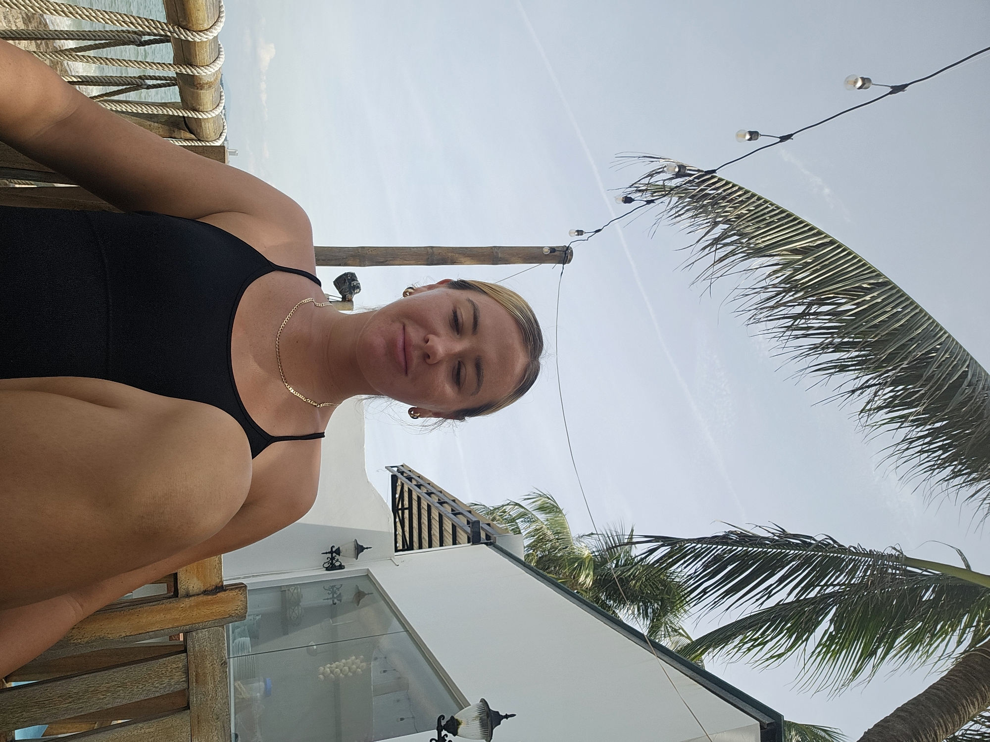 A blonde woman with closed eyes and a slight smile is sitting outdoors in Phu Thuy Ward, Vietnam. She is wearing a black tank top and a gold necklace. She is seated on a wooden chair, with her legs crossed. In the background, there is a white building with windows, palm trees, and string lights. A wooden railing with ropes is visible to her left, and beyond that, the ocean. The sky is partly cloudy.