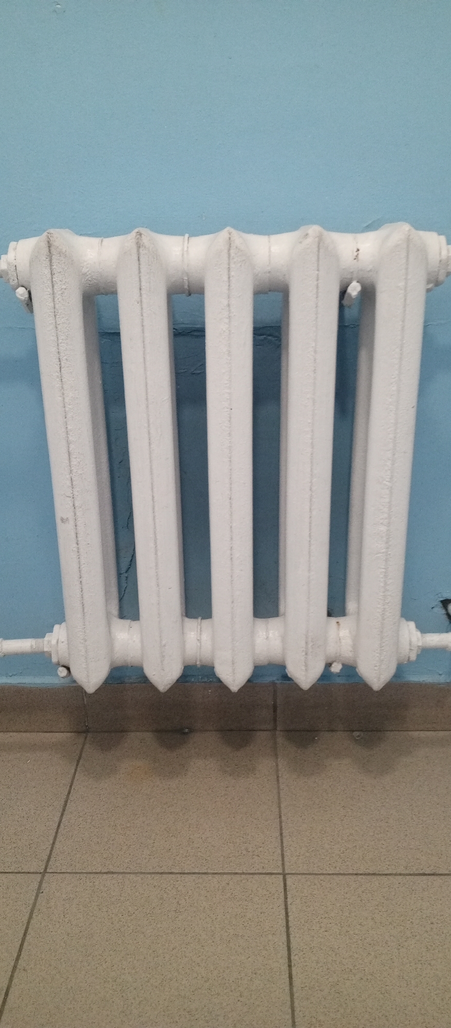 The image shows a white radiator mounted on a light blue wall. The radiator has six vertical sections and appears to be made of cast iron. It is connected to a white pipe on the left side. Below the radiator, there is a tiled floor with beige square tiles. The wall has some slight imperfections and a darker line running horizontally behind the radiator. The scene appears to be indoors, possibly in a hallway or room. There are no people visible in the image. The photograph is taken from a straight-on perspective. The location of Yeruda, Russia is not indicated in the visual elements of the image.