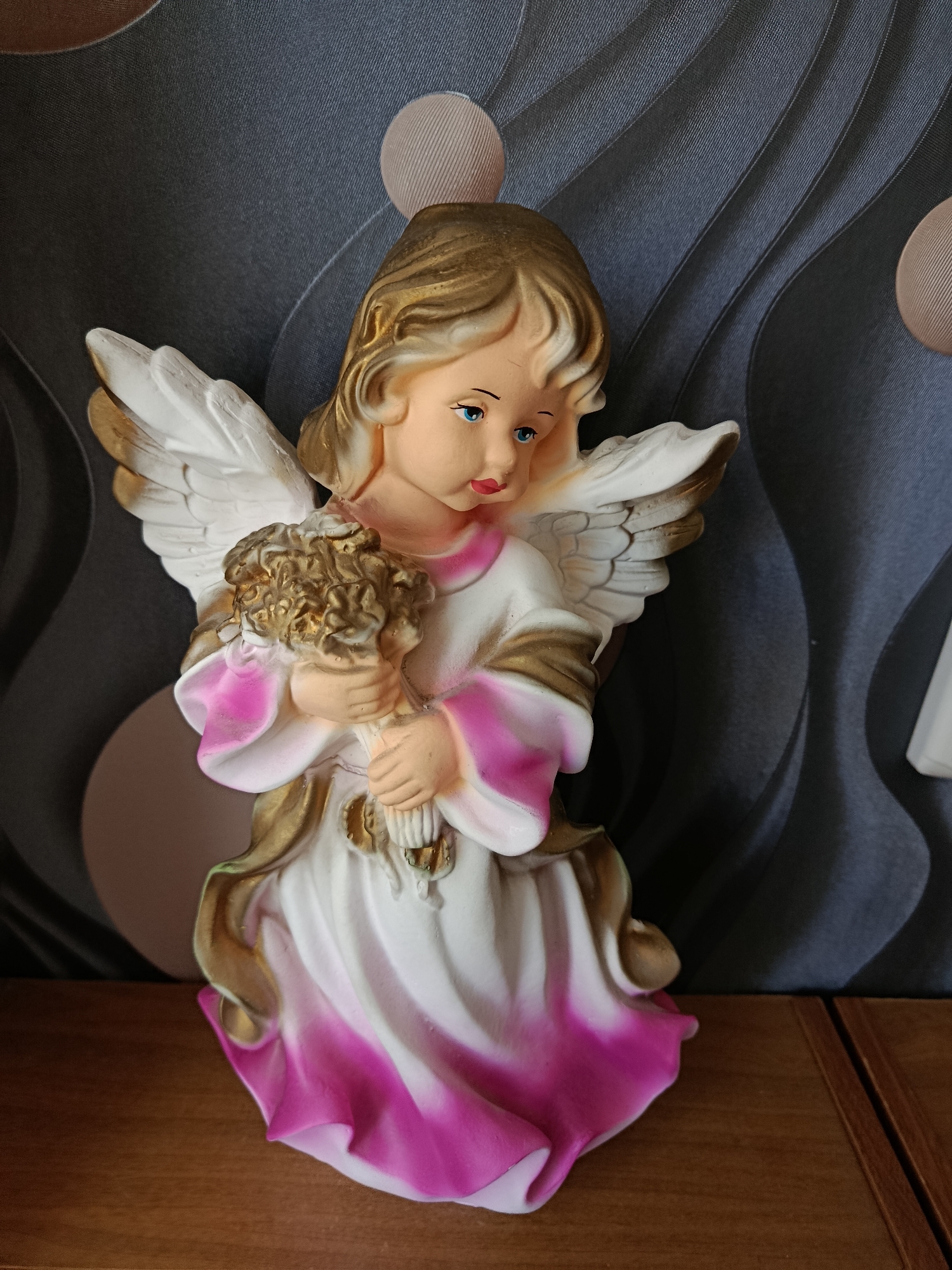 A close-up, eye-level, medium shot captures an angel figurine placed on a wooden surface in front of a textured gray and beige background with swirling patterns. The figurine, approximately 12 inches tall, is made of painted ceramic and depicts a young angel with golden hair, blue eyes, and rosy cheeks. The angel wears a white gown with pink and gold accents, and holds a bouquet of golden flowers. The angel's wings are white with subtle gold detailing. The background consists of alternating vertical stripes of dark gray and lighter gray with a wavy texture, punctuated by circular shapes of beige and lighter gray. The wooden surface appears to be a table or shelf, with visible wood grain. The lighting is soft and natural, creating subtle shadows on the figurine and background. The scene is static and does not suggest any activity or interaction. The location in Svietlahorsk, Belarus is not discernable from the visual information provided.