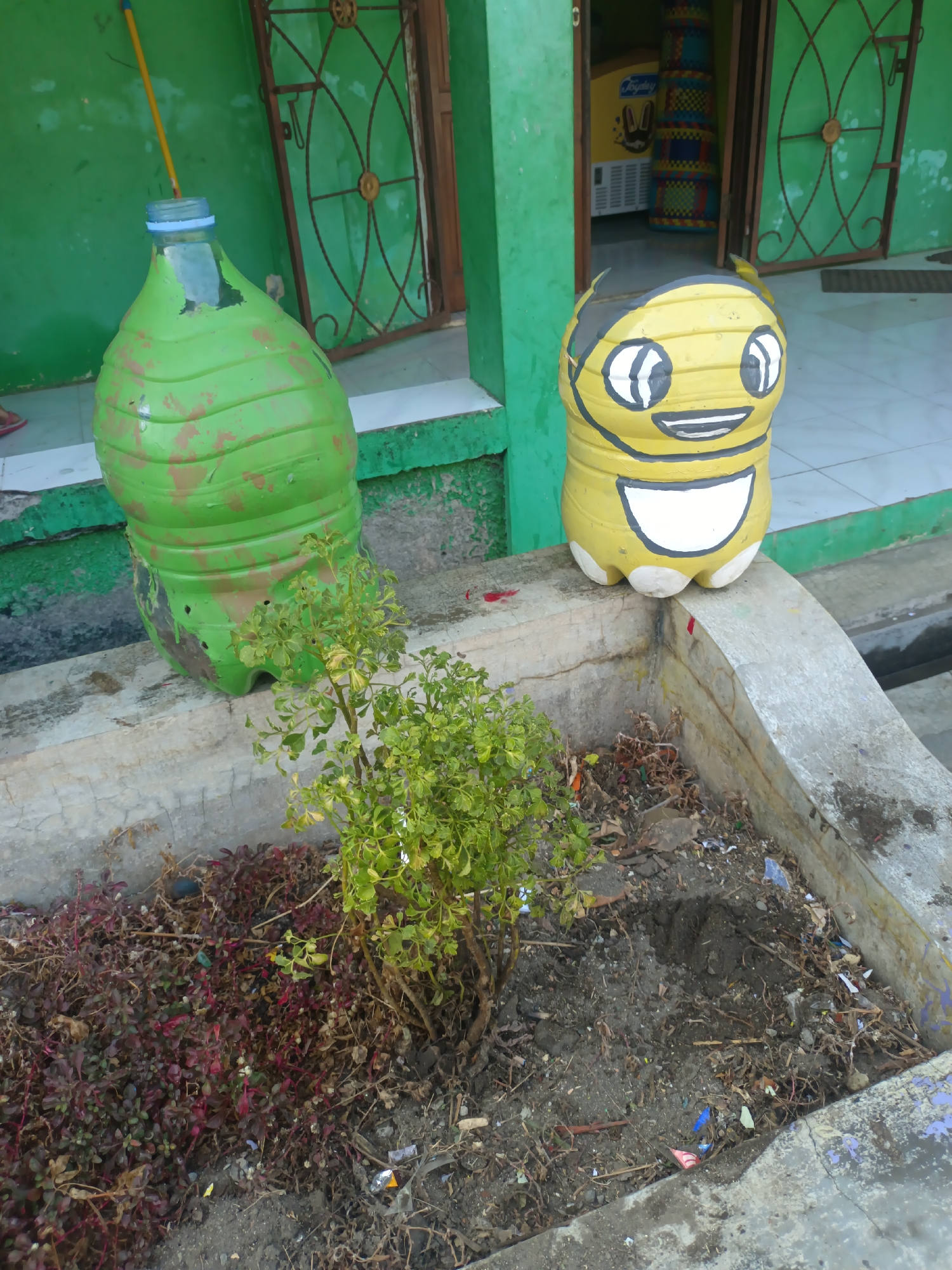 The image captures a scene in Sarimulyo, Indonesia, focusing on two repurposed plastic bottles serving as decorative planters. To the left, a large green plastic bottle is positioned, with a yellow stick protruding from its opening, possibly a plant support. The bottle shows signs of wear, with some orange-brown paint streaks over the green base coat. Adjacent to it, on the right, stands a yellow plastic bottle painted to resemble a cartoon character with large white eyes outlined in black, a wide black mouth with white teeth, and a white bib-like area. Both bottles sit on a concrete ledge. In front of the ledge is a planting bed filled with soil and a small green leafy plant. The ground surrounding the bed is littered with debris, including small pieces of plastic and dried leaves. In the background, a green building with windows featuring ornate metal grills is visible. The overall environment suggests an outdoor, possibly urban or semi-urban, setting in Sarimulyo.