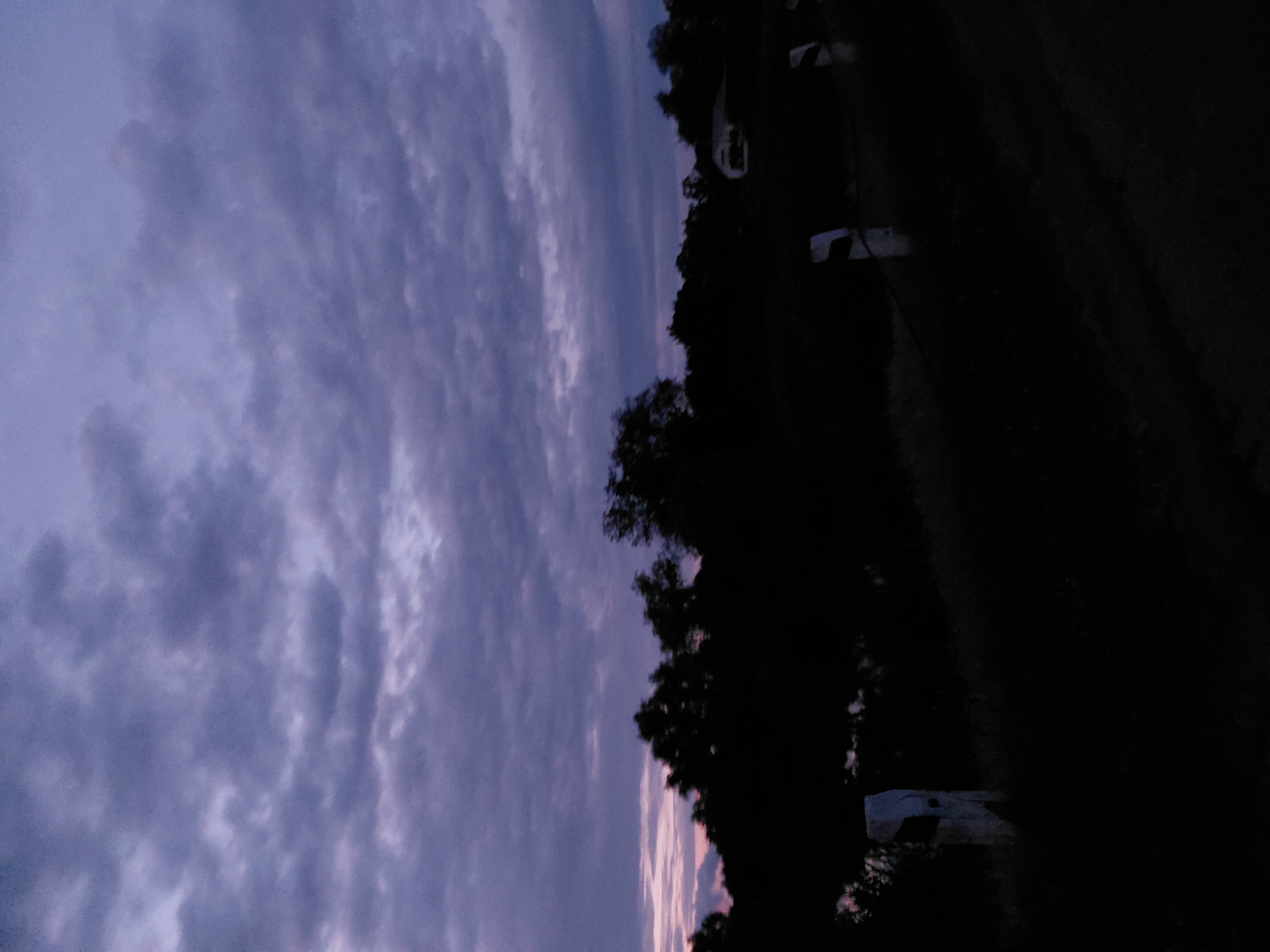 The image depicts a dimly lit, rural landscape at dusk or dawn, likely in Ustyvytsia, Ukraine. A dark, silhouetted tree line runs across the middle ground, with some lighter areas suggesting a body of water on the left and a small structure on the right. The sky above is filled with heavy, dark purple and gray clouds, with a faint streak of pink visible on the left horizon, indicative of the low light conditions. In the foreground, two white posts with black tops are visible, suggesting a roadside or boundary marker. The overall impression is one of a serene, quiet, and slightly melancholic natural setting.