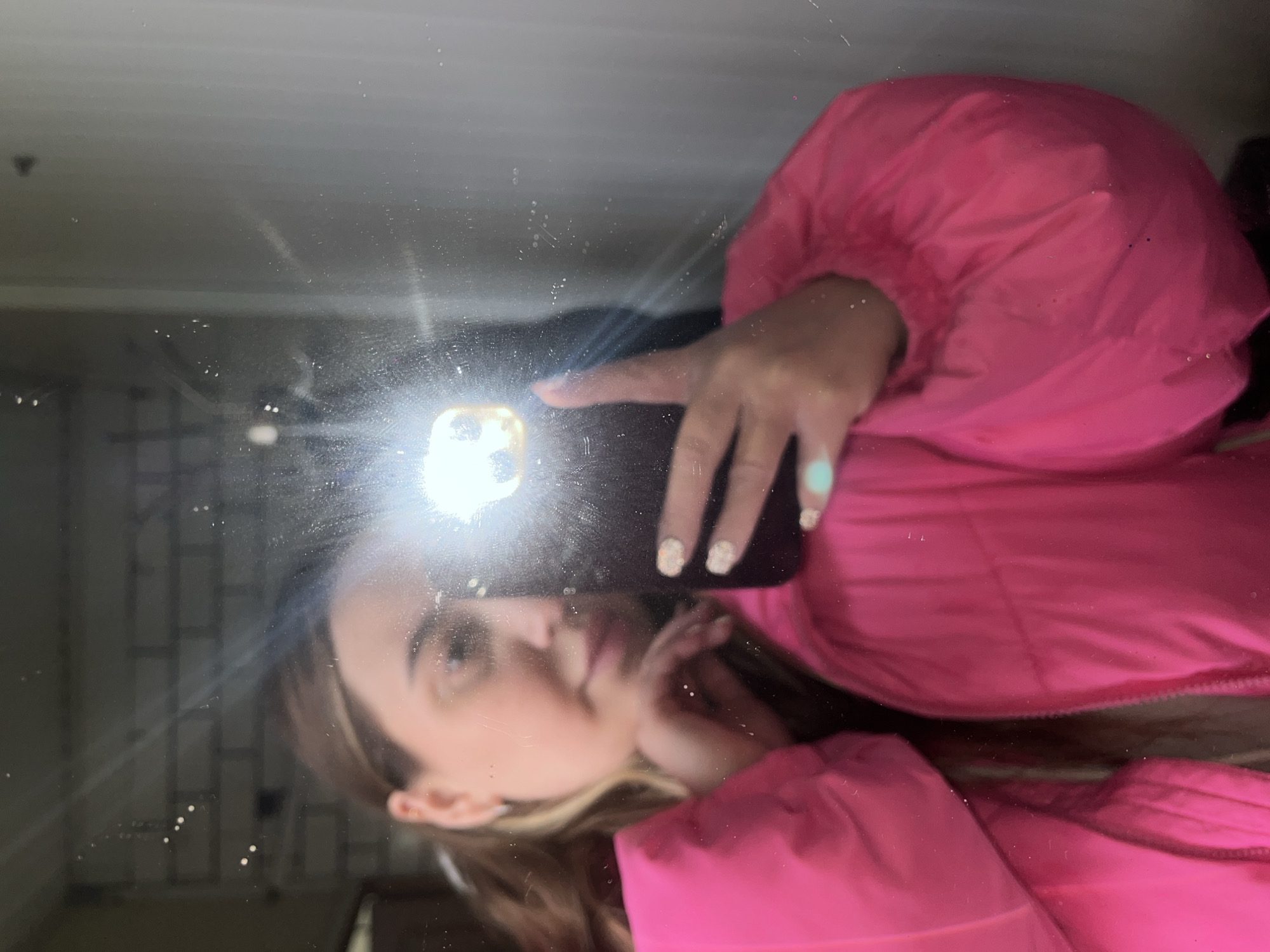 A person wearing a pink puffer jacket is taking a selfie in a mirror. The camera flash is obscuring some details. The background appears to be an interior space with a window. The location and specific details are not discernible from the image.