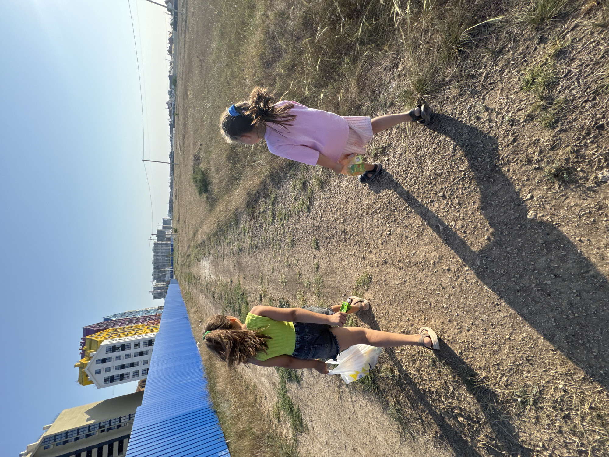 Two young girls walk away from the camera down a dirt path on a sunny day. The girl on the left wears a lime green tank top and denim shorts, carrying a white plastic bag. The girl on the right wears a pink t-shirt and light pink shorts, holding a plastic bag with bottles. In the background, a bright blue fence runs parallel to the path, separating it from construction sites with tall buildings under a clear blue sky. The surrounding area is dry, grassy terrain with scattered bushes.